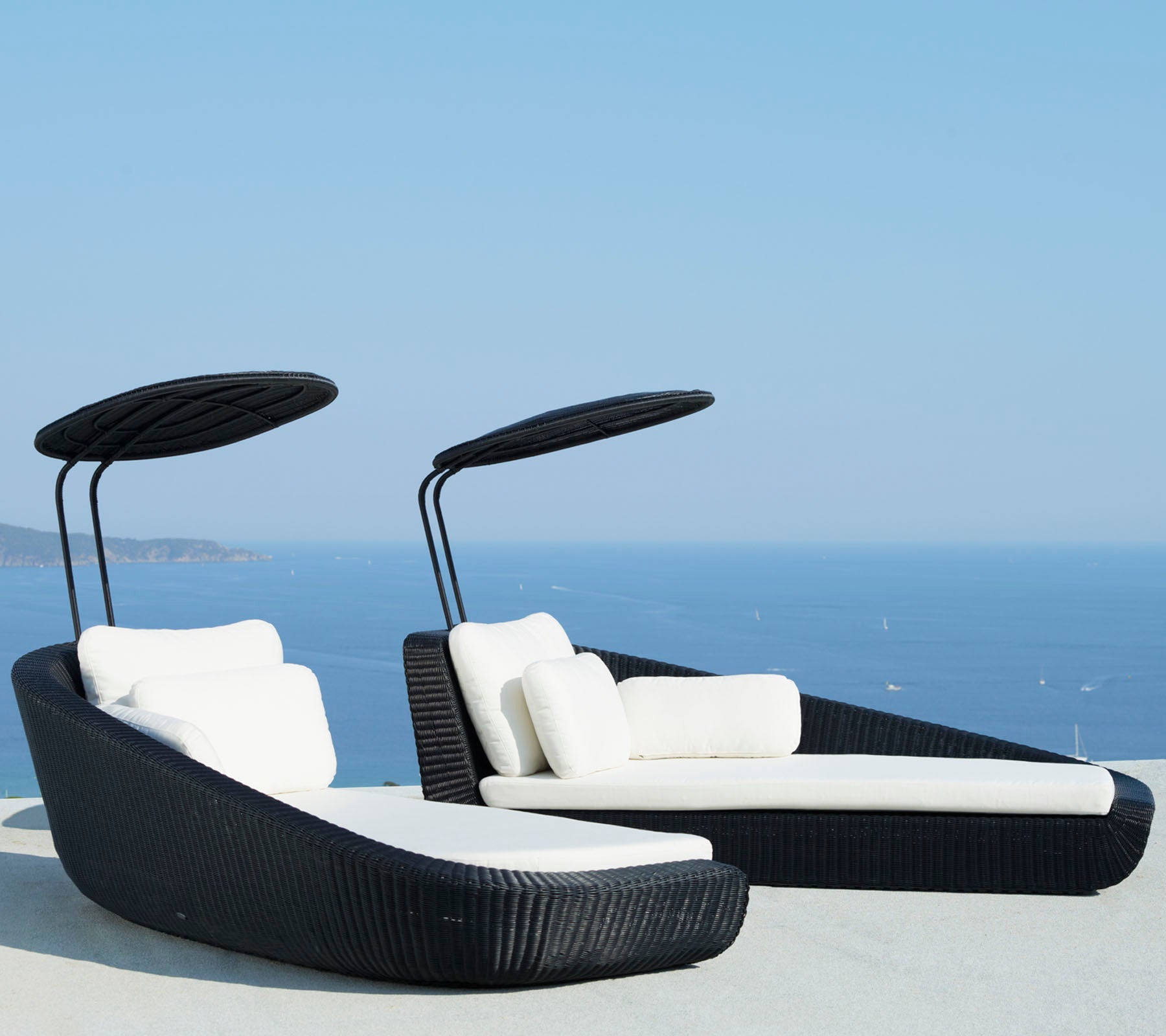 Twee stijlvolle lounge stoelen met witte kussens en gebogen luifels, tegen een schilderachtige blauwe oceaanachtergrond.