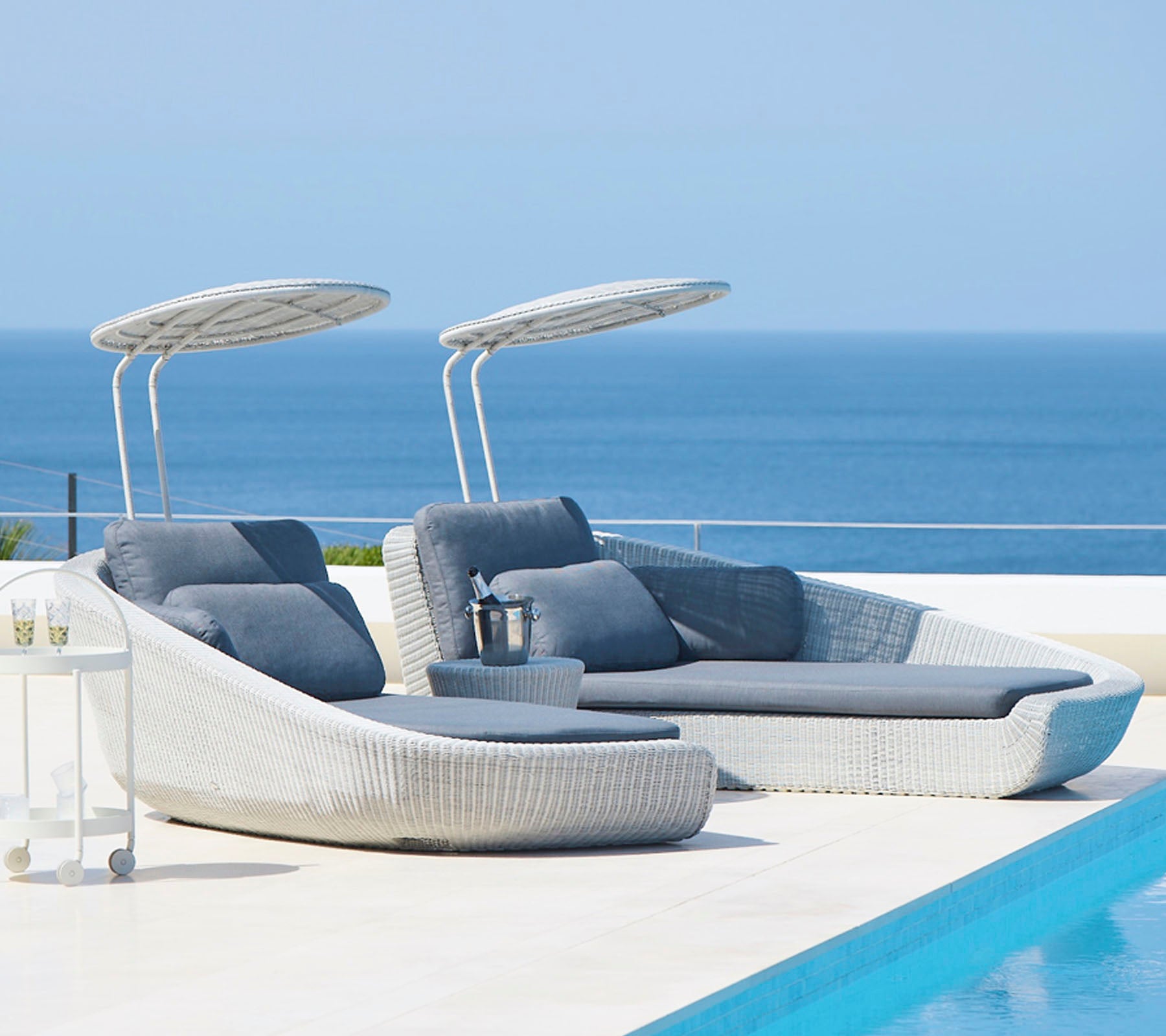 Stijlvolle ligstoelen voor buiten met overkappingen naast een serene pool, met uitzicht op een kalme zee. Perfect voor ontspanning en vrije tijd.