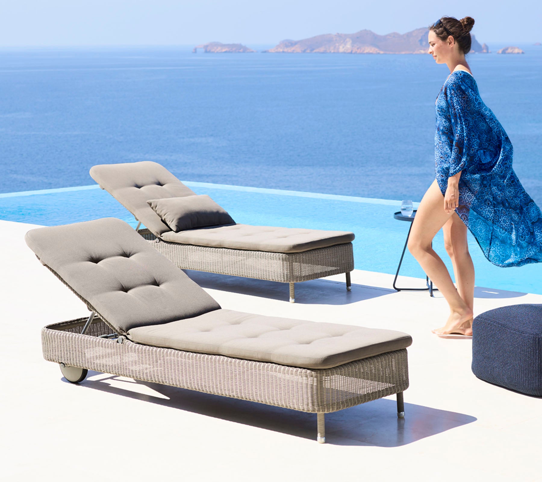 Een vrouw in een vloeiende blauwe cover-up loopt naast twee stijlvolle ligstoelen bij een zwembad met uitzicht op een serene oceaan.