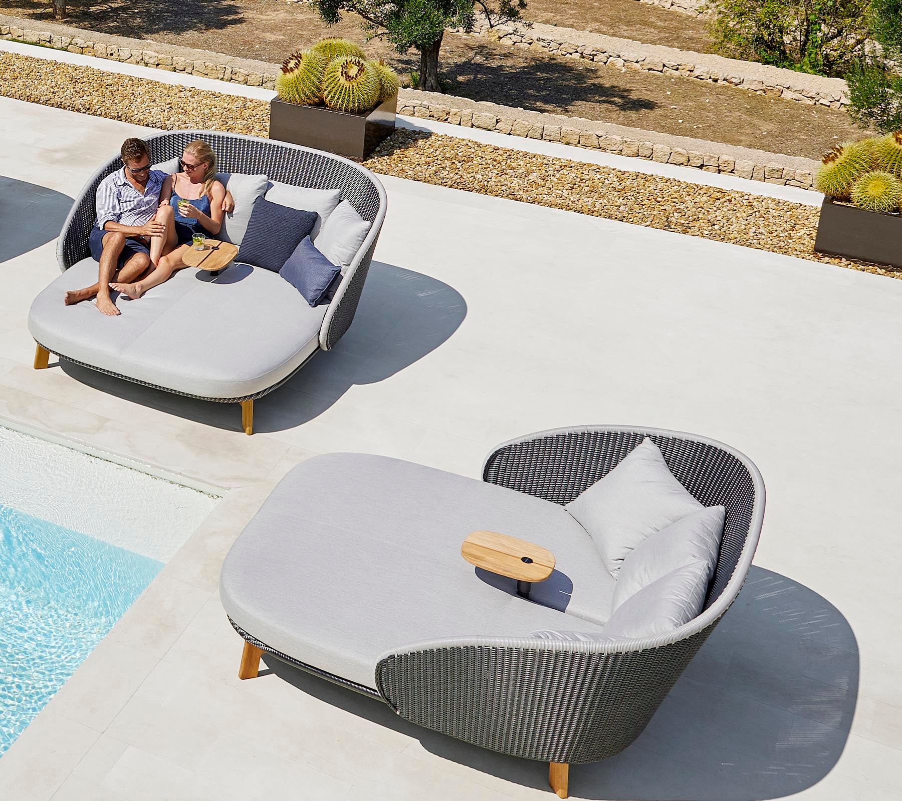 Twee mensen ontspannen op stijlvolle ligstoelen buiten bij een zwembad en genieten van hun vrije tijd met kussens en een tafeltje.