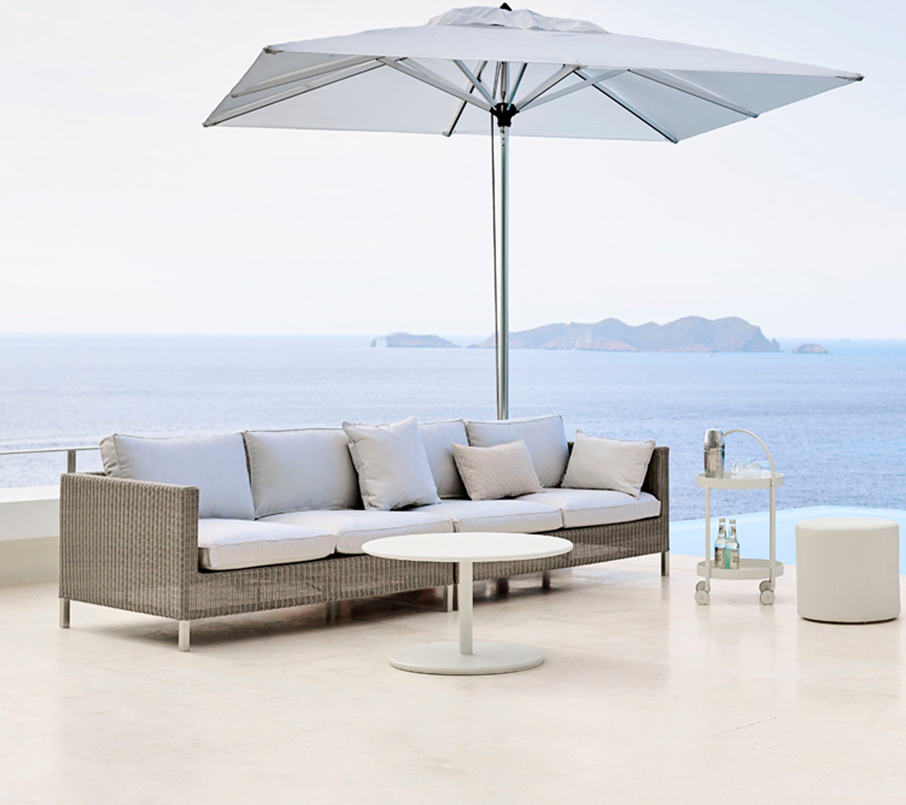 Stijlvol buitenzitgedeelte met een lichtgrijze sofa, ronde tafel en een groot parasol, met uitzicht op een sereen wateroppervlak.