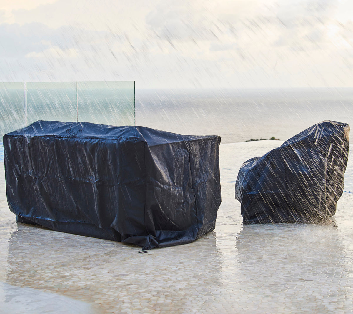 Tuinfurnitur bedekt met donkere beschermhoezen, beschermd tegen regen, met een achtergrond van een bewolkte lucht en de oceaan.
