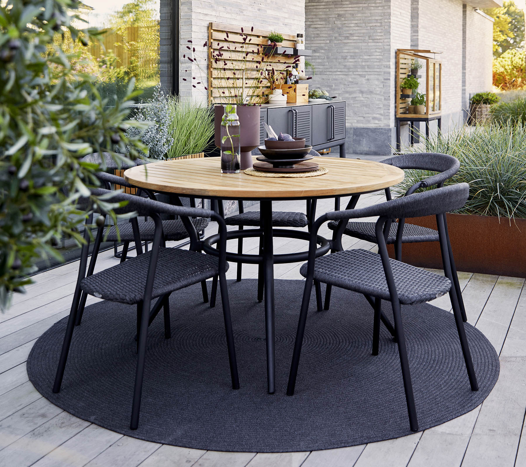 Rond houten tafel met zwarte stoelen eromheen, gedekt voor het diner, omgeven door groen en een gestructureerd tapijt.