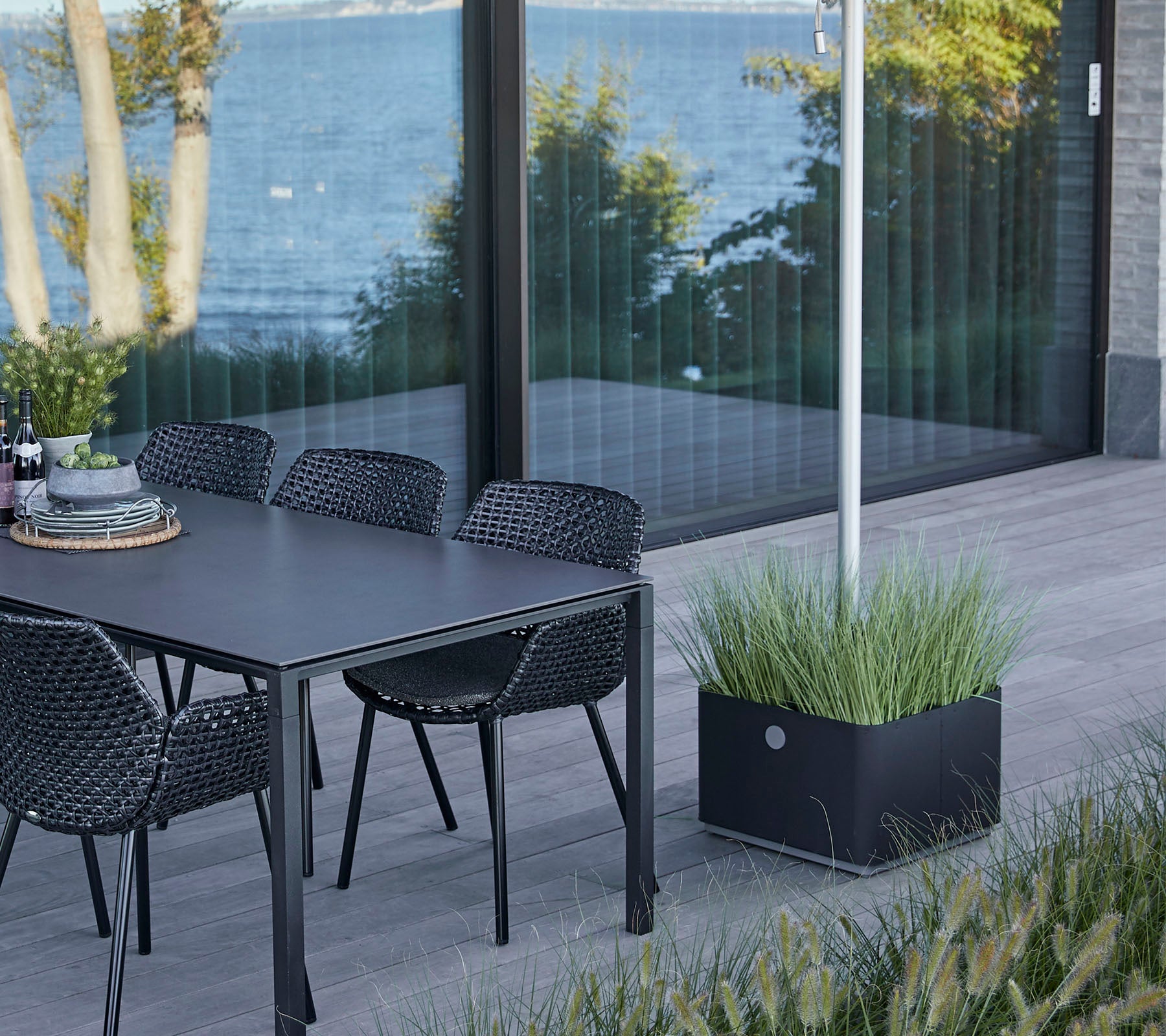 Een stijlvolle tafel met stoelen naast een uitzicht op het water, met een decoratieve plantenbak met weelderig gras.