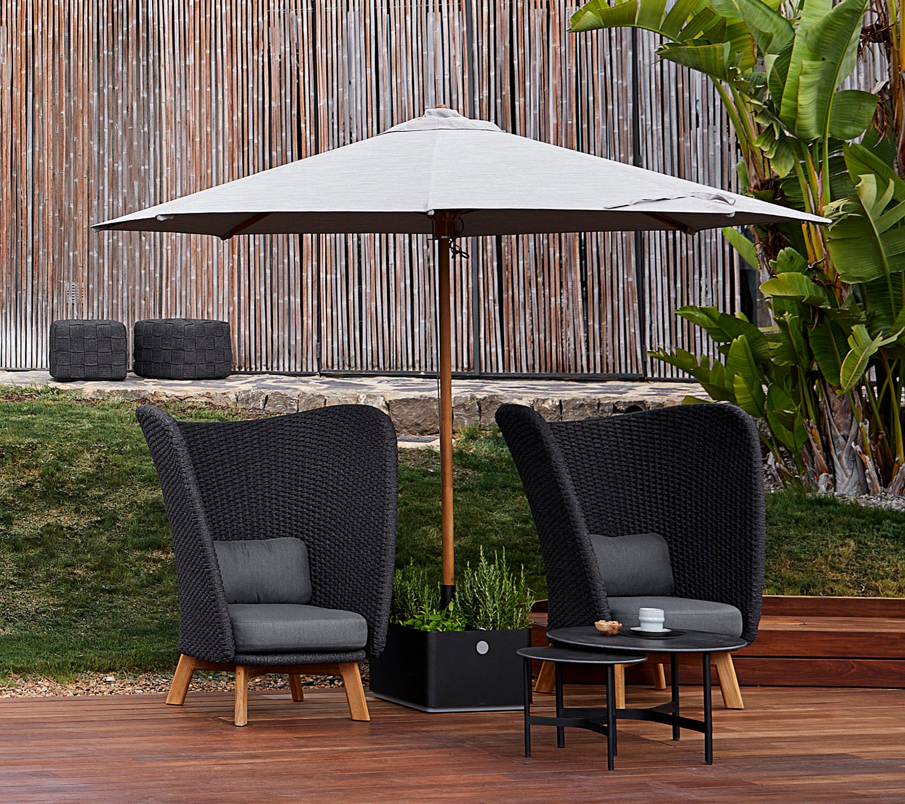 Twee stijlvolle zwarte stoelen met grijze kussens en een klein tafeltje onder een grote parasol, omgeven door groen en houten vloeren.