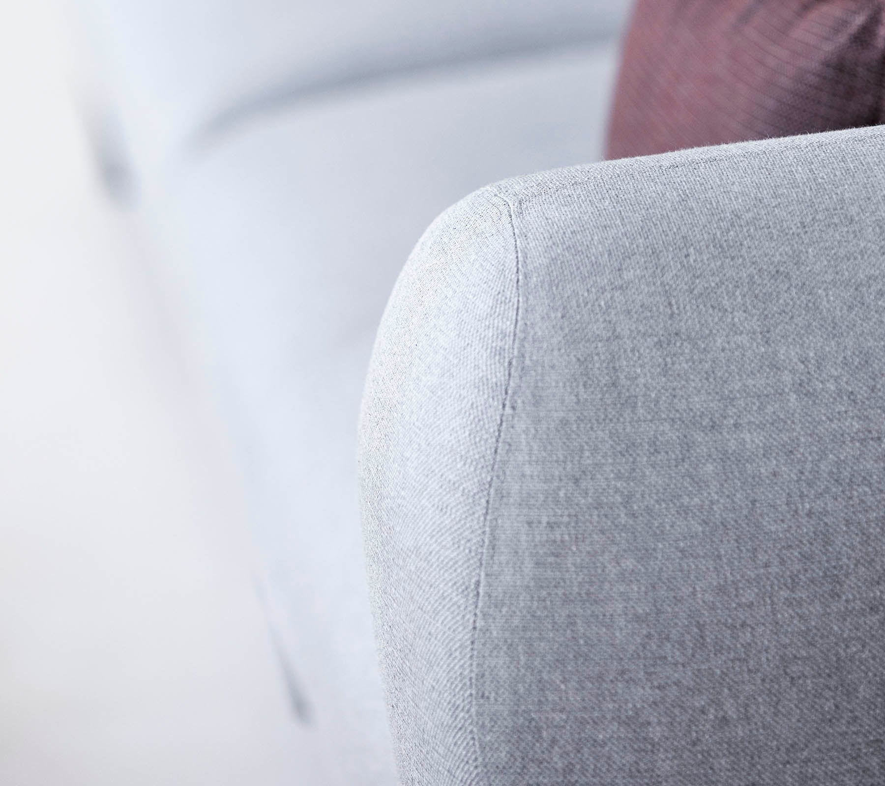 Close-up van een lichtgrijze sofa-arm, met een gestructureerde stof en een zacht, afgerond ontwerp, met een hint van een donker kussen op de achtergrond.