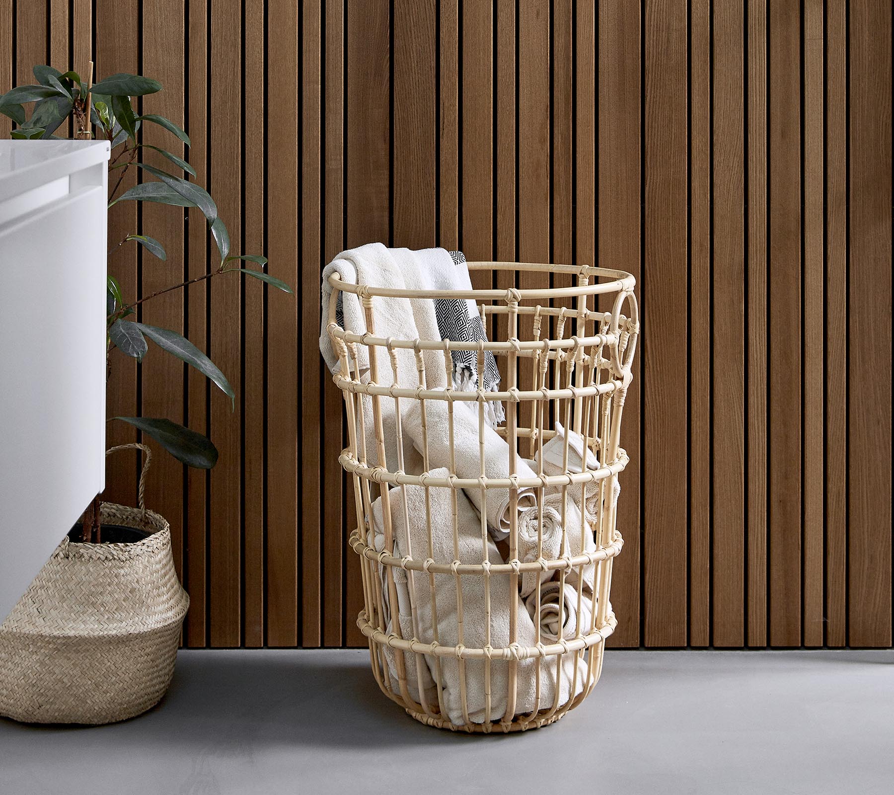 Geweven wasmand gevuld met opgerolde handdoeken, geplaatst naast een plant en een gestructureerde houten muur.
