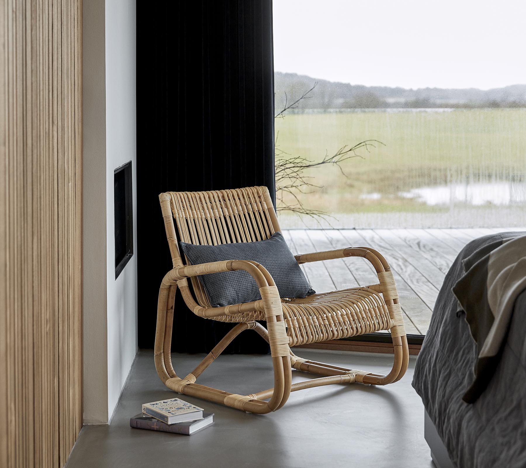 Een stijlvolle schommelstoel gemaakt van natuurlijke materialen, geplaatst naast een bed en een boek, met een schilderachtig uitzicht zichtbaar door grote ramen.