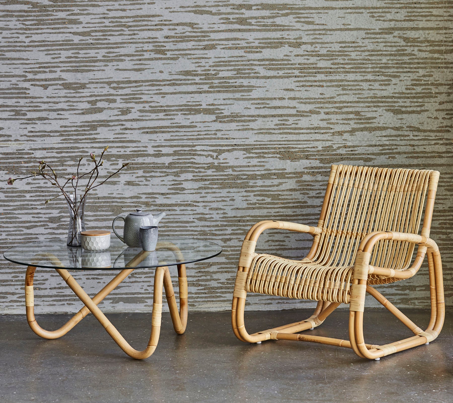 Rattannoord en glazen tafel met decoratieve elementen, geplaatst tegen een textuurmuur. Een minimalistische en moderne decoratiestijl.