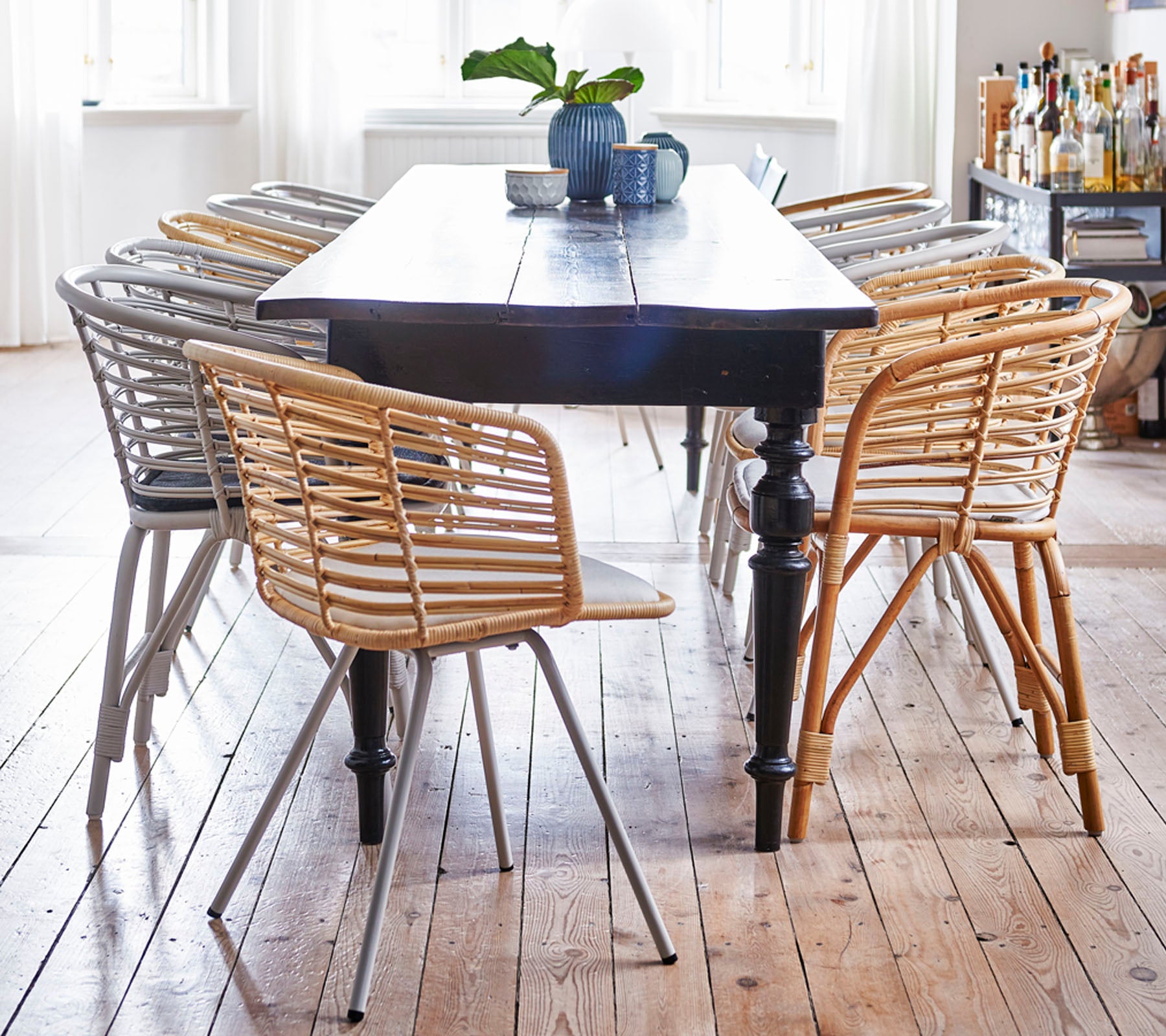 Een houten tafel omringd door stijlvolle, geweven stoelen in lichte en donkere tinten, met een kleine plant en een mok bovenop.