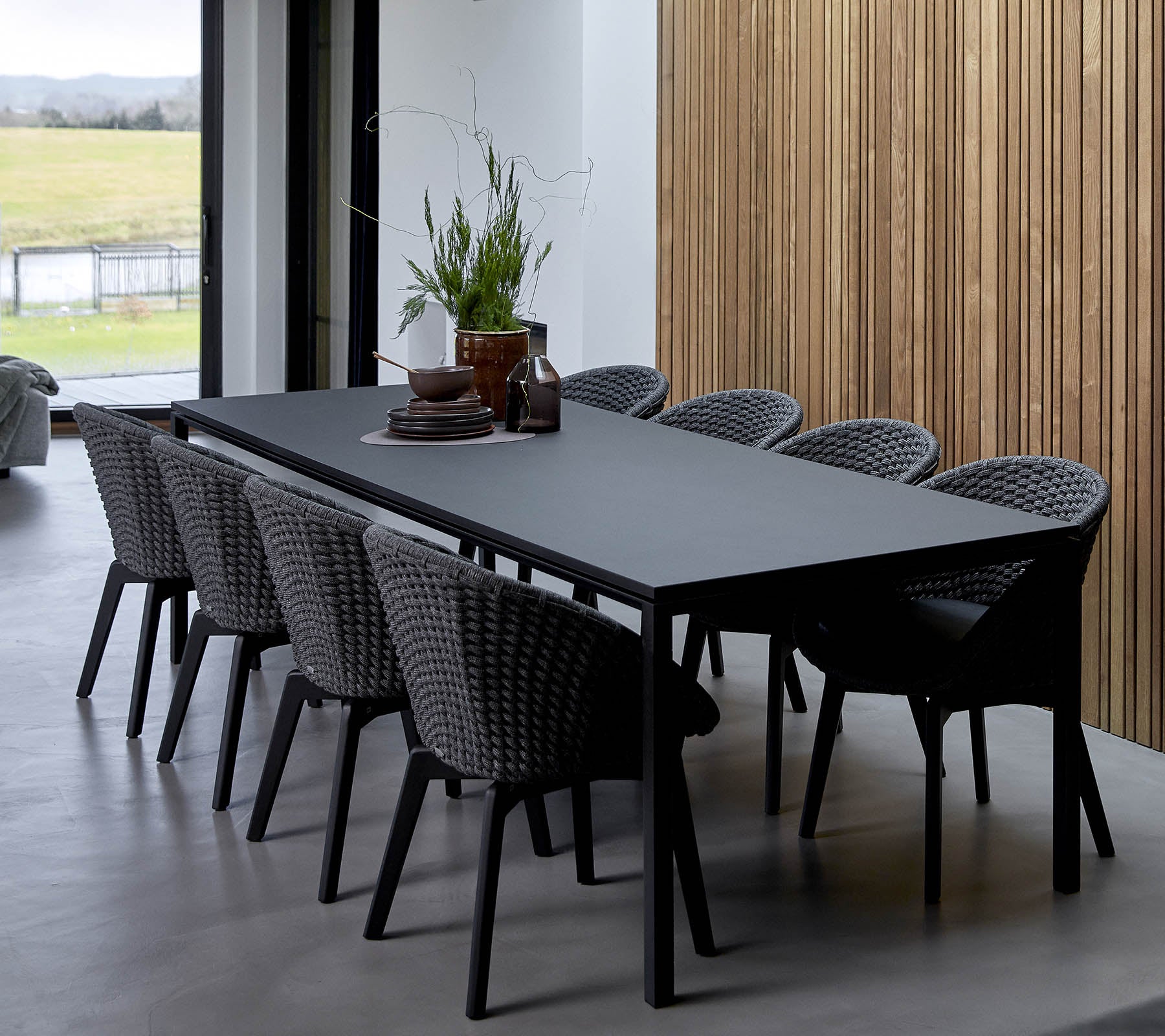 Een gestroomlijnde tafel omringd door geweven stoelen, versierd met decoratieve planten en een middelpunt, tegen een houten achtergrond.