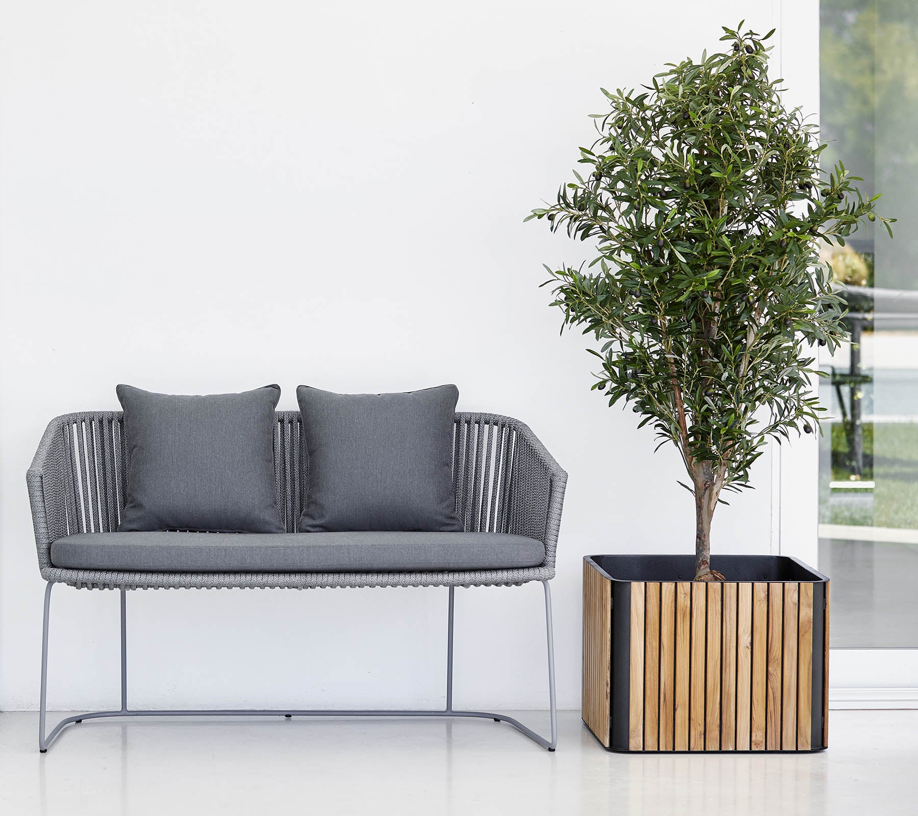 Moderne bank met twee kussens naast een potplant in een decoratieve bloempot, tegen een schone, neutrale achtergrond.