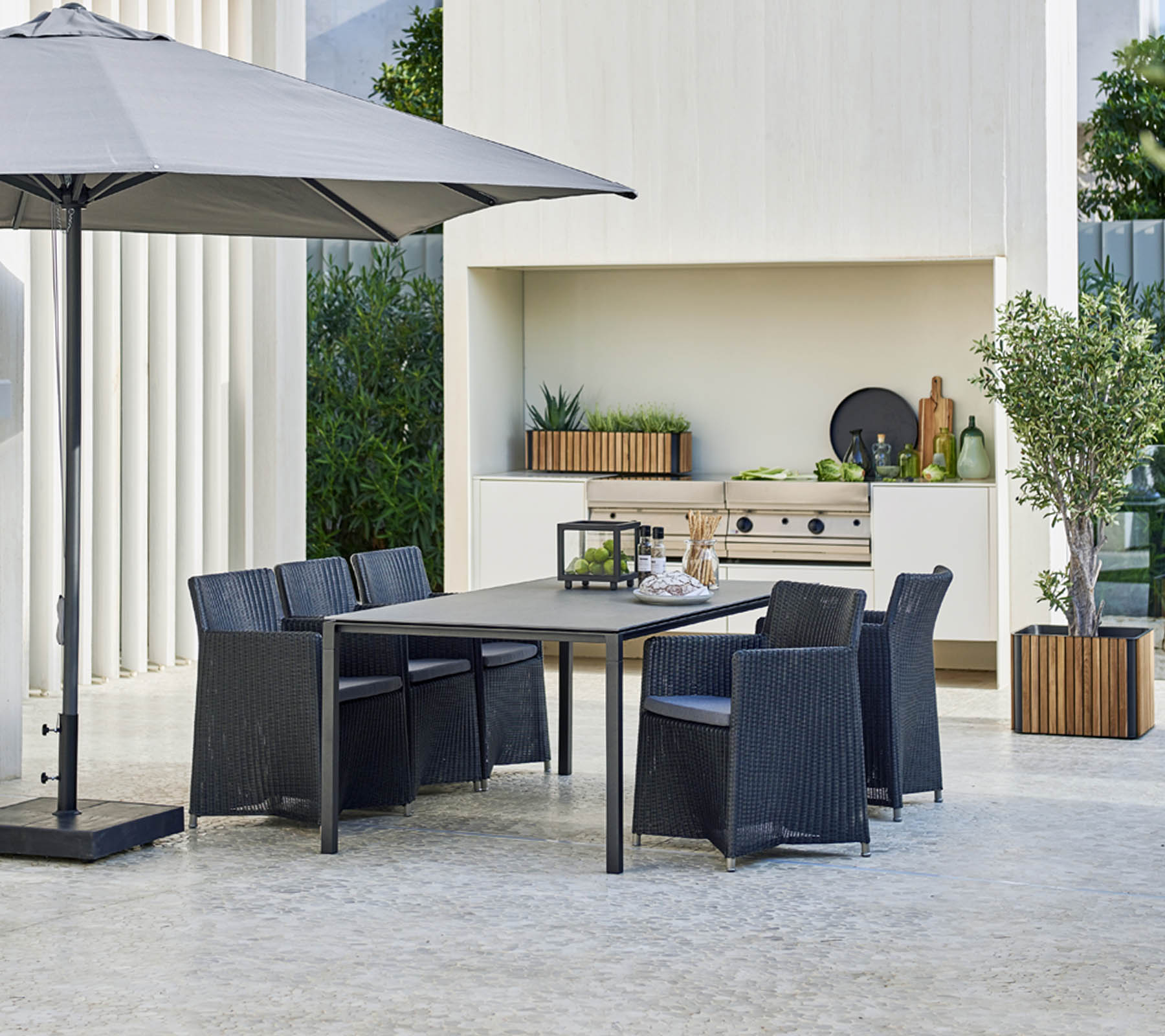 Moderne buitentafelset met een grote tafel, zes stoelen, een stijlvolle parasol en een achtergrond met groen en decoratieve elementen.