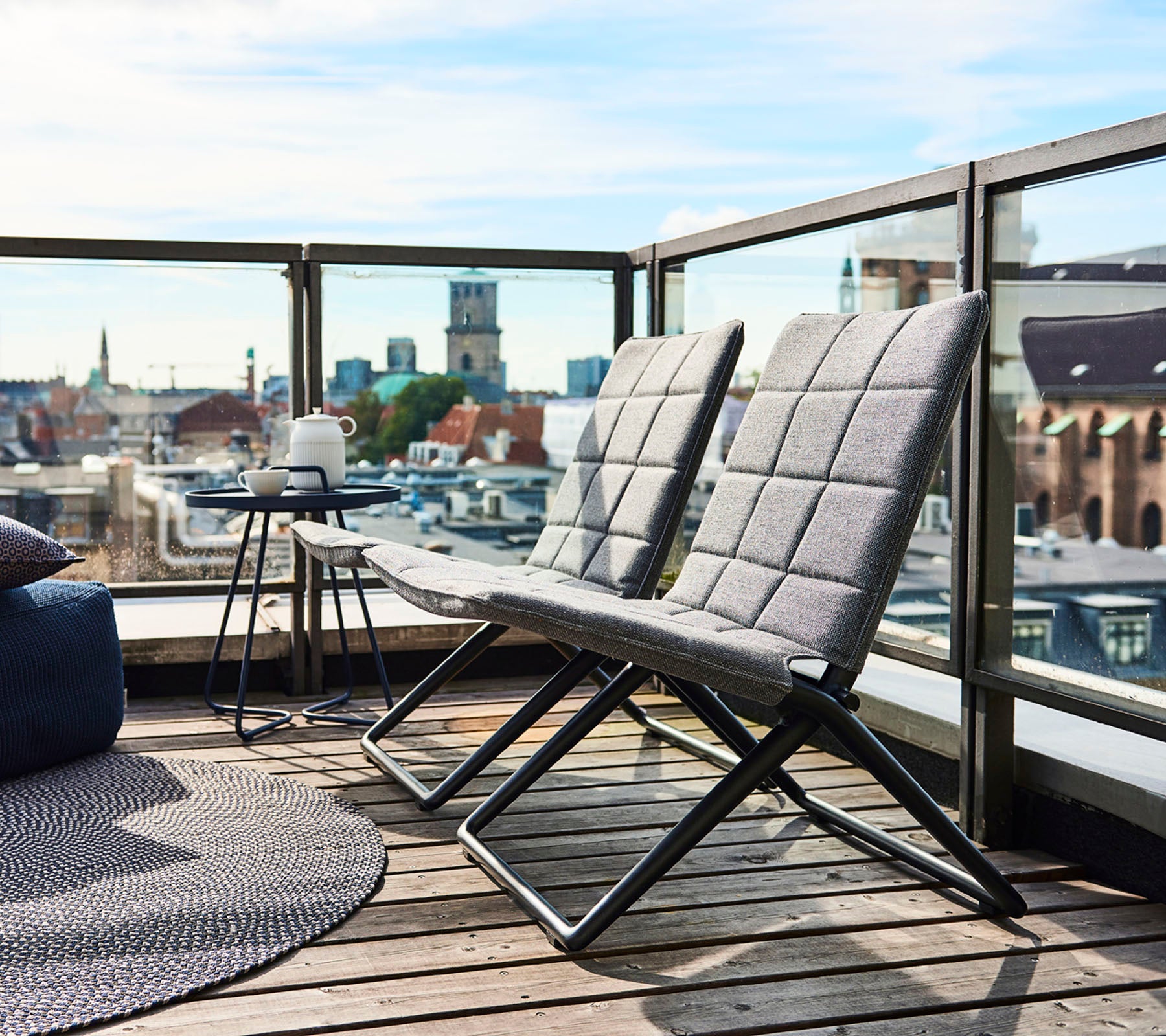 Twee moderne lounge stoelen met slanke frames en gestructureerde kussens, vergezeld van een klein tafeltje aan de zijkant en uitzicht op de stad op de achtergrond.