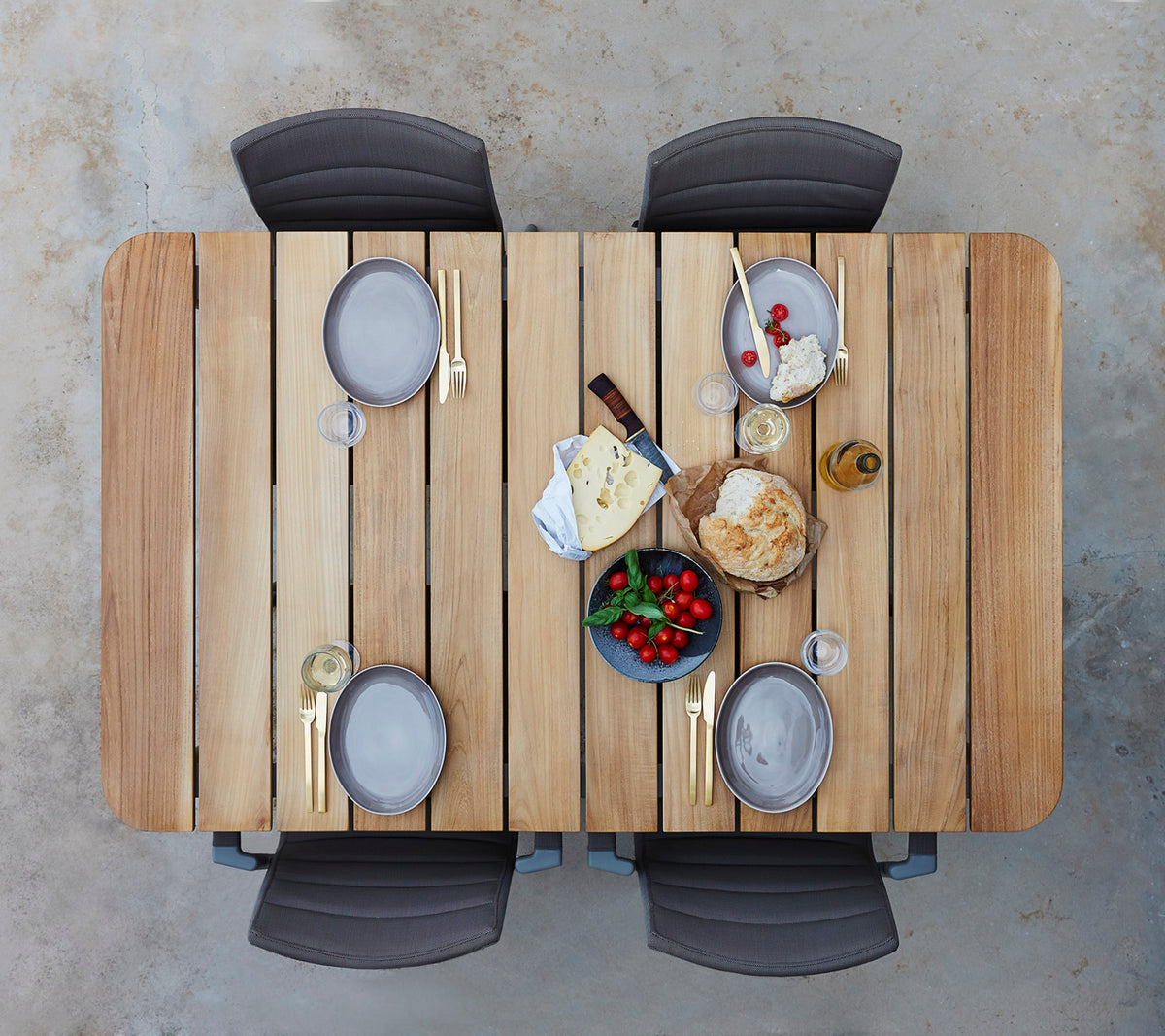 Een houten tafel gedekt met borden, glazen en een assortiment van kazen, fruit en brood, omringd door vier stoelen.