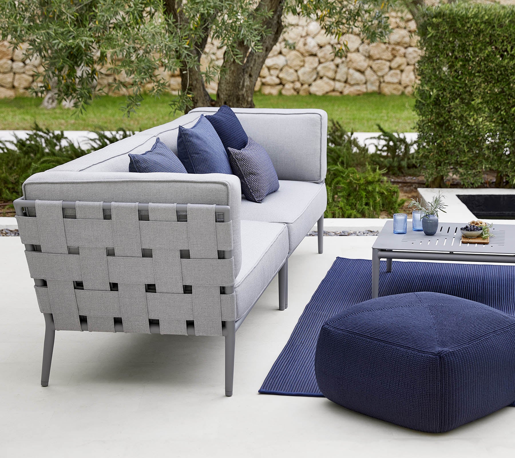 Moderne buitensitgelegenheid met een grijze bank met decoratieve kussens, een stijlvolle salontafel en een gezellige pouf.