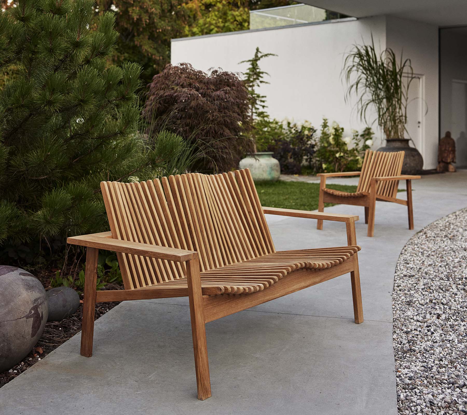 Twee houten Amaze-loungestoelen en een bijpassende bank hebben een lattenontwerp, omringd door groen en decoratieve elementen.