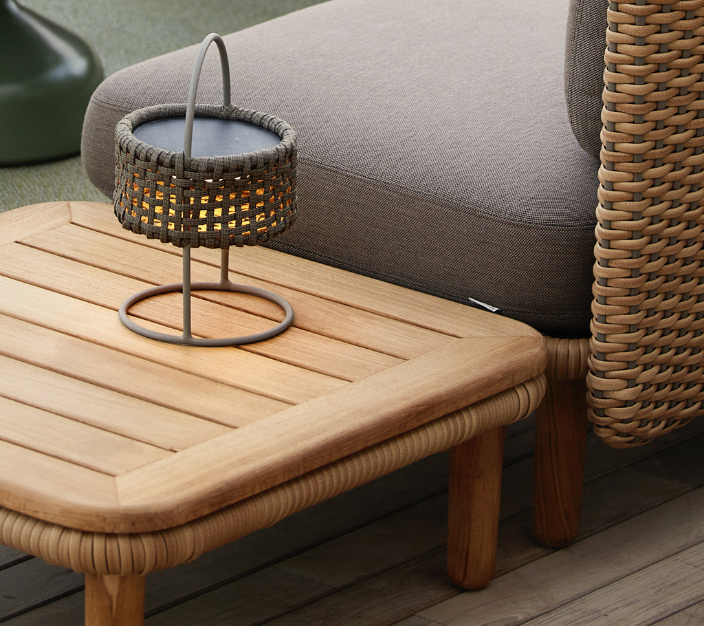 Teakhouten salontafel met een lamellenblad, vergezeld van een geweven lamp, die een gezellige en uitnodigende sfeer creëert.