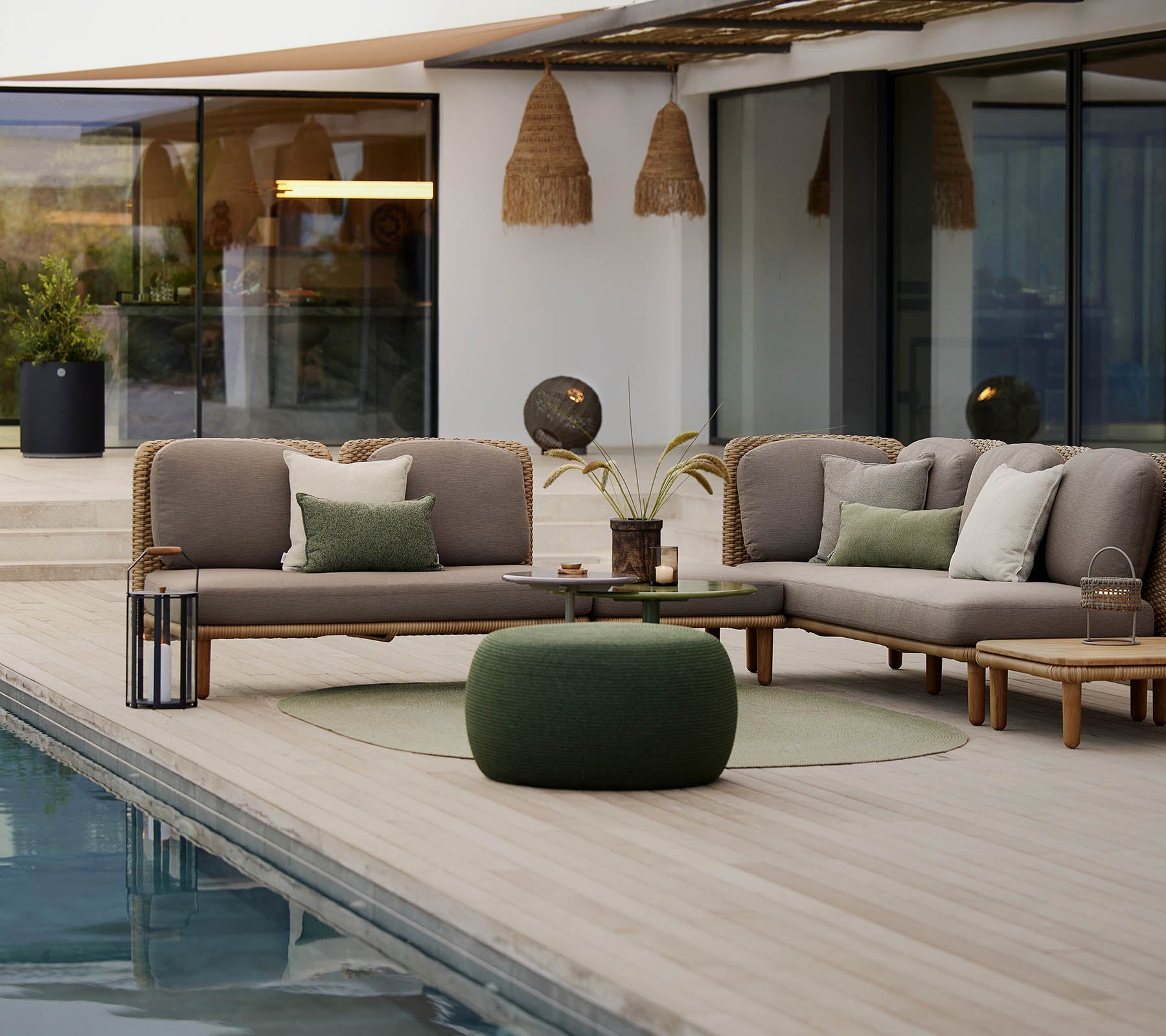 Een stijlvolle loungehoek met een sectionele bank met kussens, een ronde tafel en een gezellige pouf, naast een rustige pool.