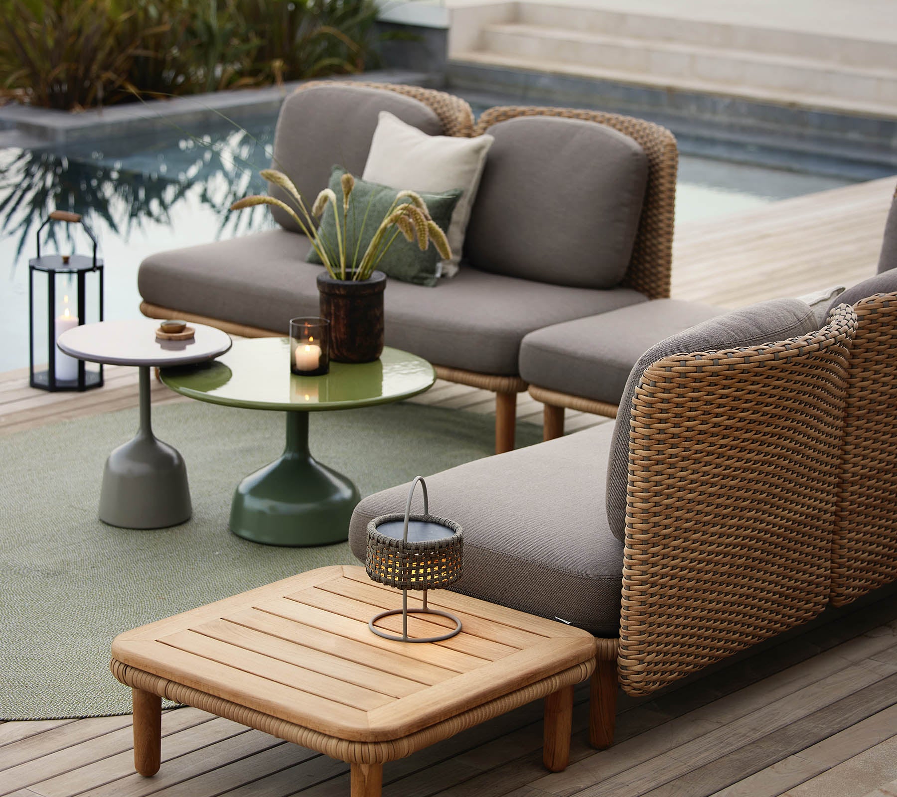 Een stijlvolle lounge zithoek met een grijze sofa met rotan accenten, een groene bijzettafel en decoratieve accessoires.