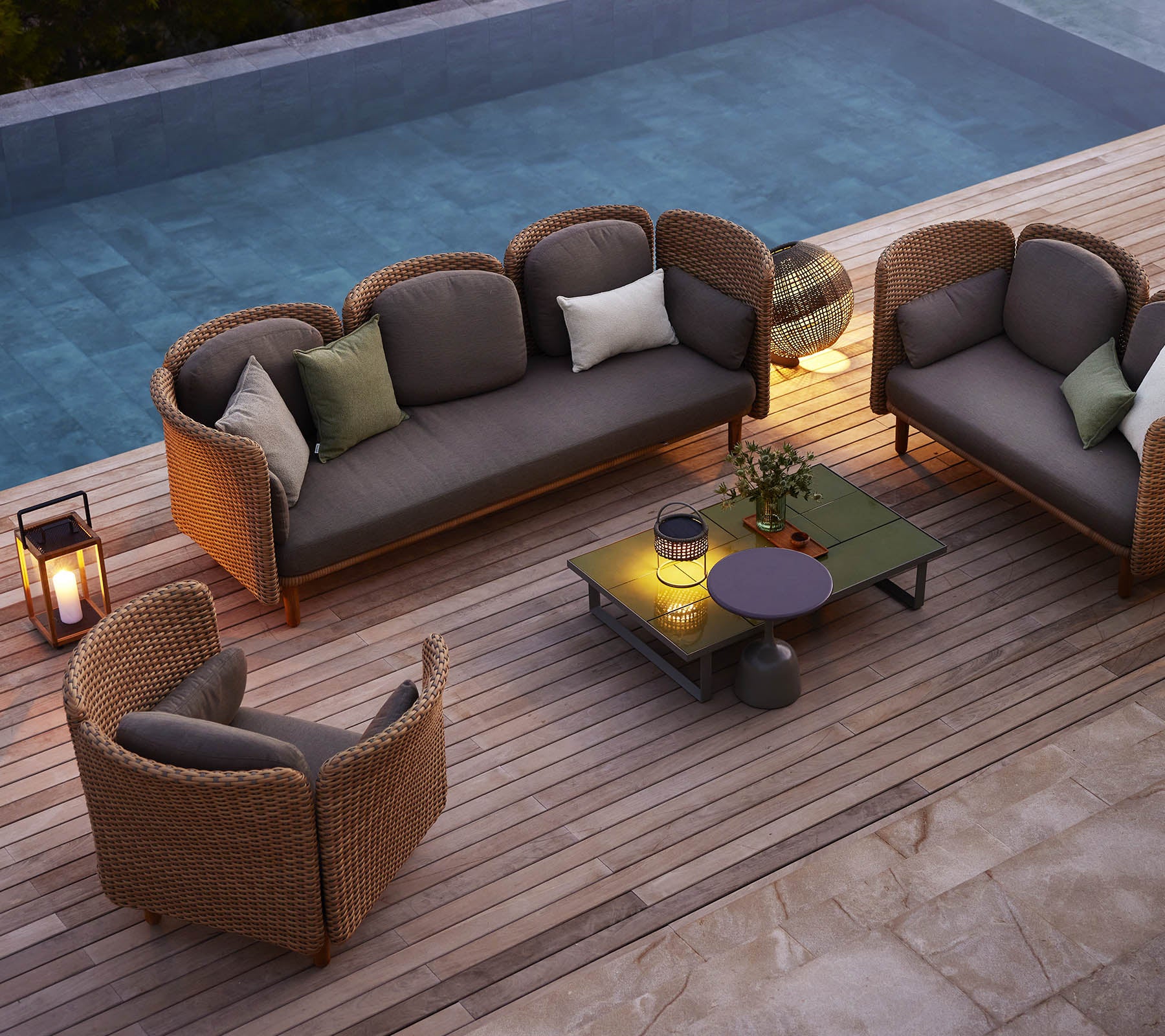 Een gezellige zitarrangement met een bank en stoelen met kussens, omringd door groen en een zwembad, op een houten terras.