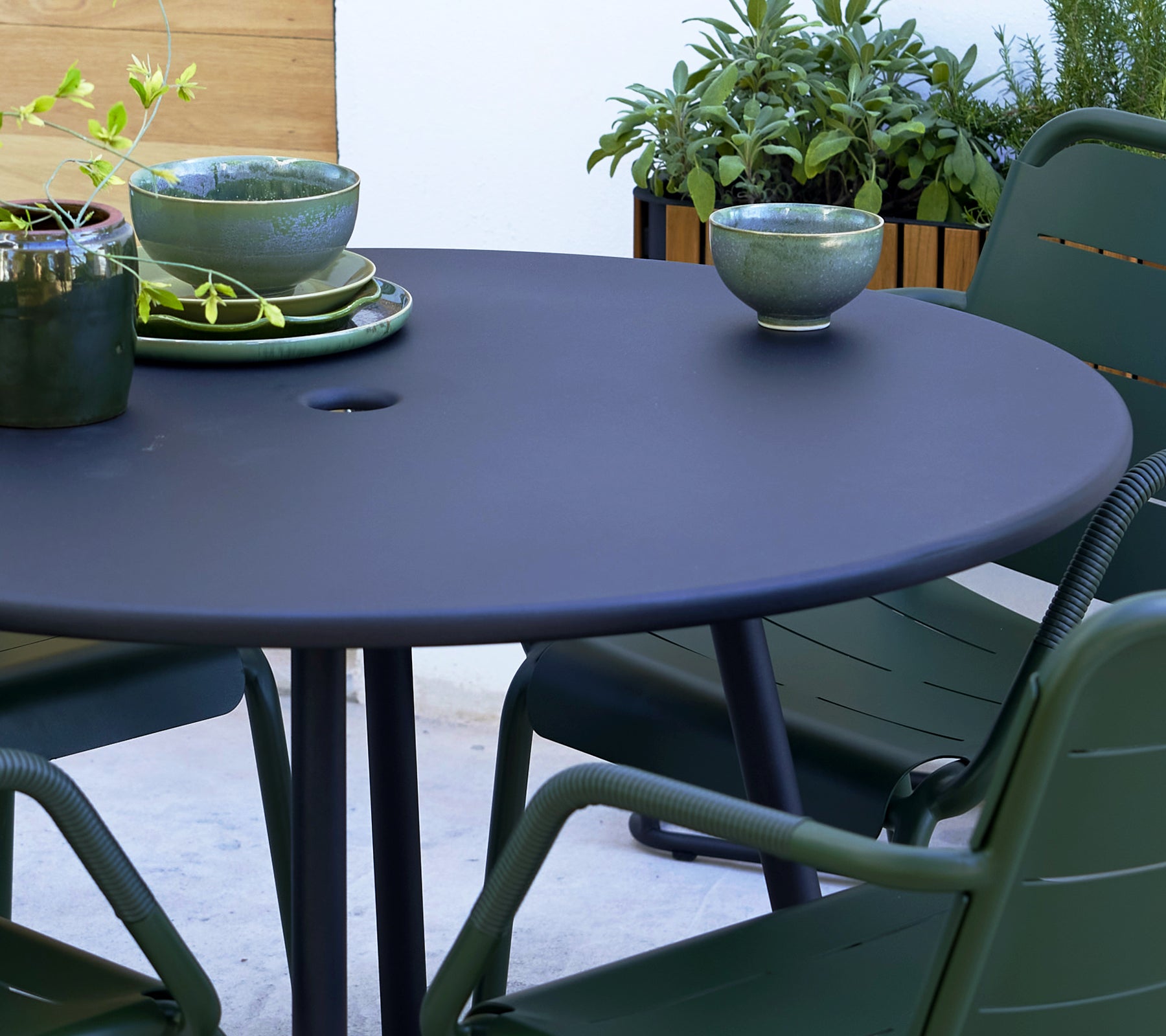 Ronde grijze tafel met groene Kopenhagen-stoelen, met decoratieve kommen en groen op de achtergrond.