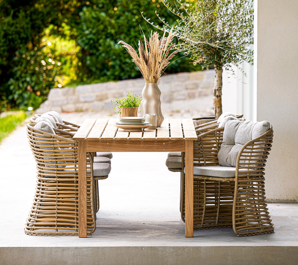 Een houten eettafel met stijlvolle rieten stoelen en een decoratieve vaas met gedroogde bloemen, aangevuld met groen in de achtergrond.