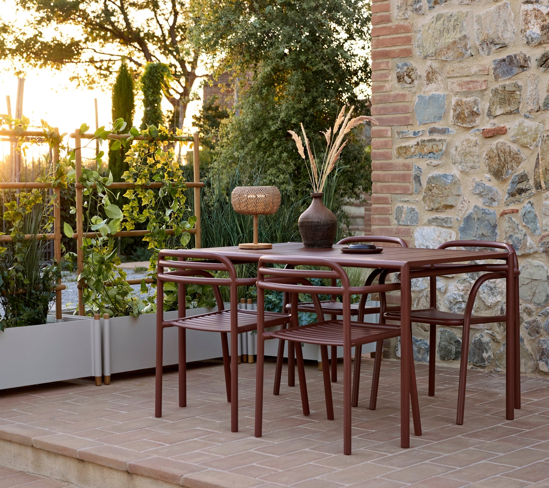 Buitenopstelling met een Bliss-tafel met stoelen, een Bliss-stoel en een Sticks-plantenbak omringd door groen bij zonsondergang.