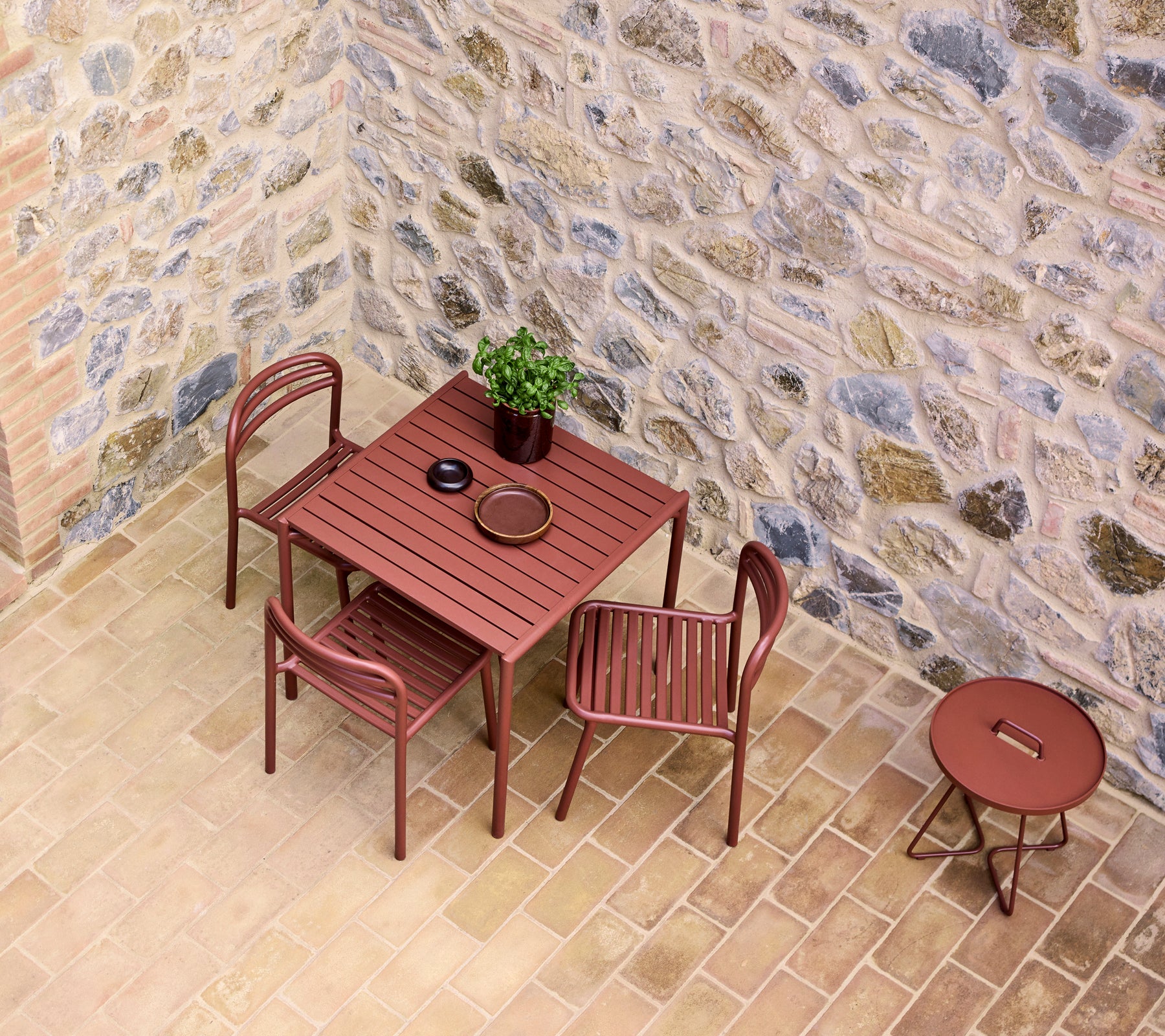 Een tafel en stoelen in een woestijnrode tint, naast een kleine ronde tafel, tegen een gestructureerde stenen muur met een potplant.