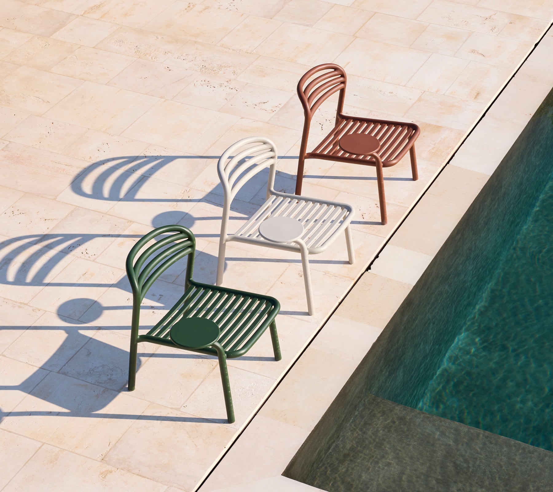 Drie moderne stoelen in groen, wit en bruin naast een zwembad.