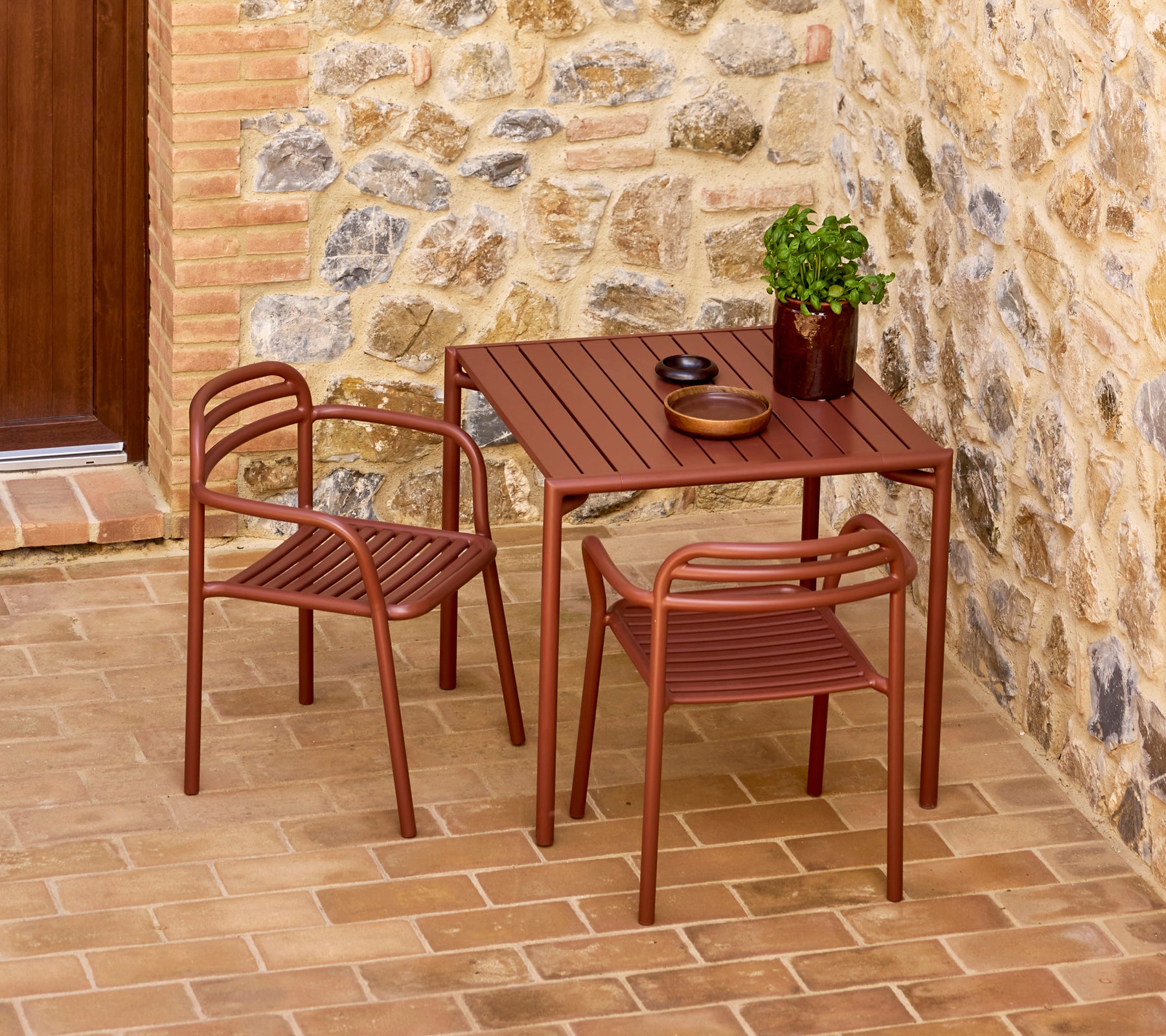 Stylish woestijnrood Bliss-tafel met twee bijpassende stoelen, vergezeld van een decoratieve plant en eenvoudig servies tegen een stenen achtergrond.