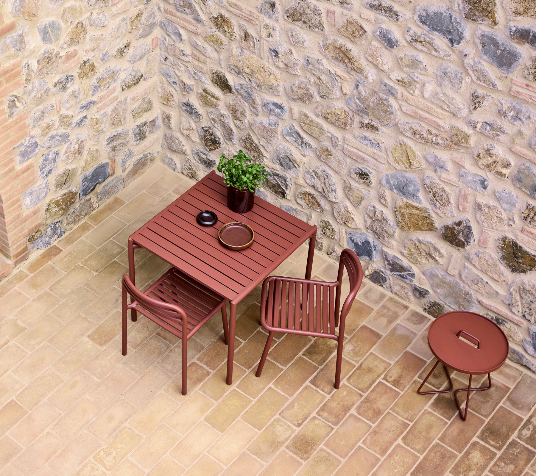een rode café tafel gezet met twee stoelen, met een kleine plant en een bord, tegen een stenen muur.