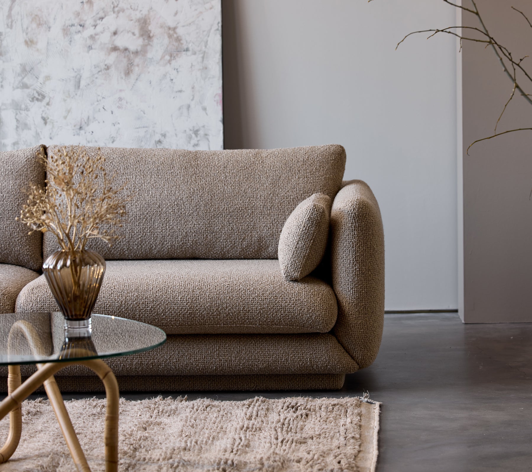 Een taupekleurige salontafel met een glazen blad staat naast een textuurbeige bank versierd met een decoratieve vaas en gedroogde bloemen.