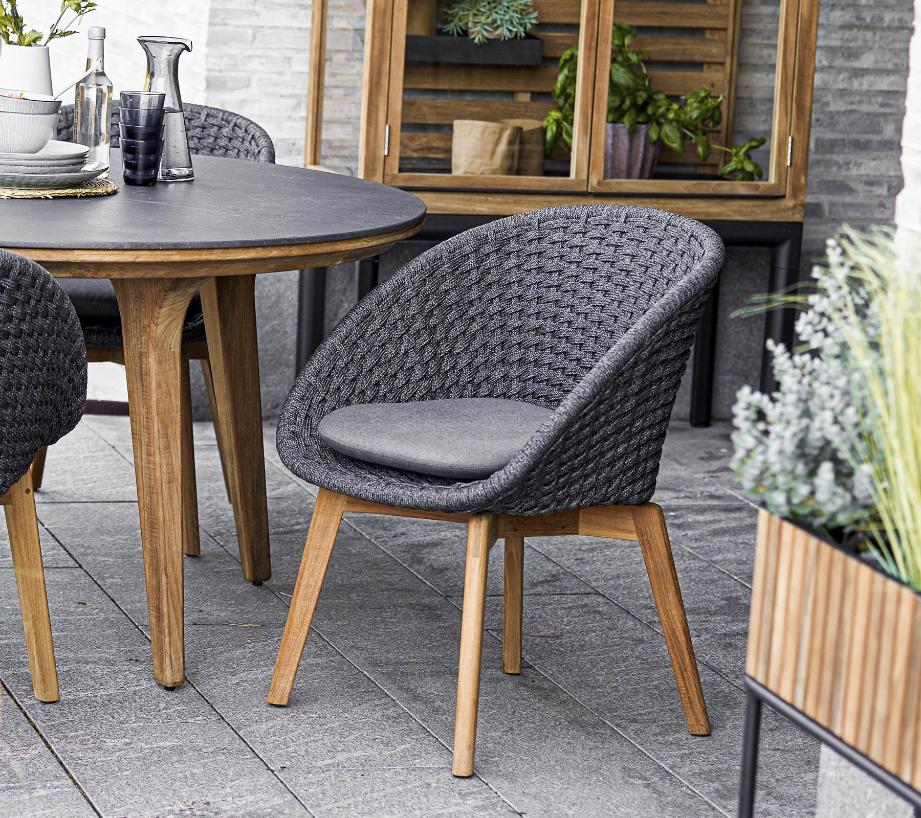 Stijlvolle geweven stoel met een gewatteerde zitting, gecombineerd met een ronde tafel, omringd door groen in plantenbakken. Perfect voor sociale bijeenkomsten.