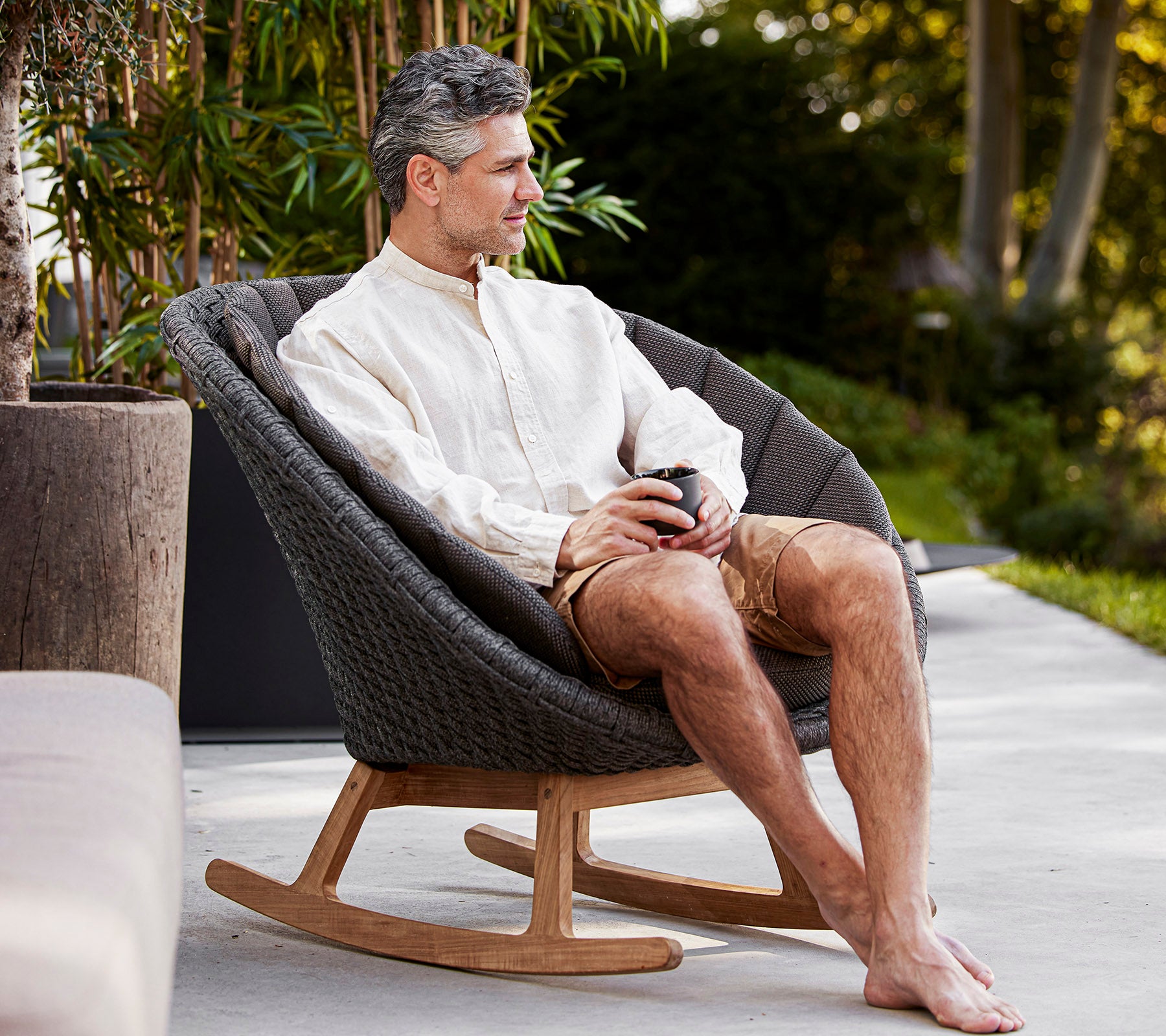 Een man zit comfortabel in een stijlvolle schommelstoel, geniet van een drankje in een rustige omgeving, casual gekleed in een licht shirt en shorts.
