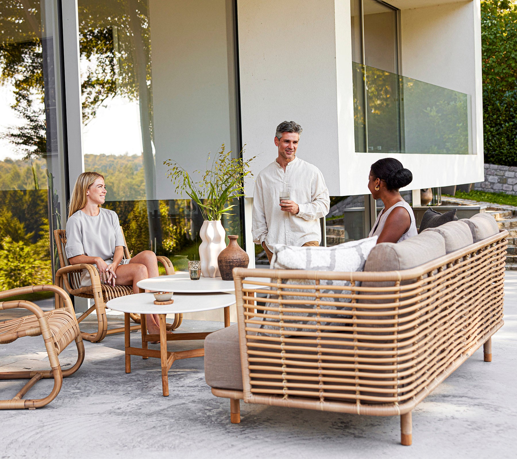 Drie mensen genieten van een informele conversatie op stijlvolle rattan meubels met decoratieve elementen, omringd door groen.