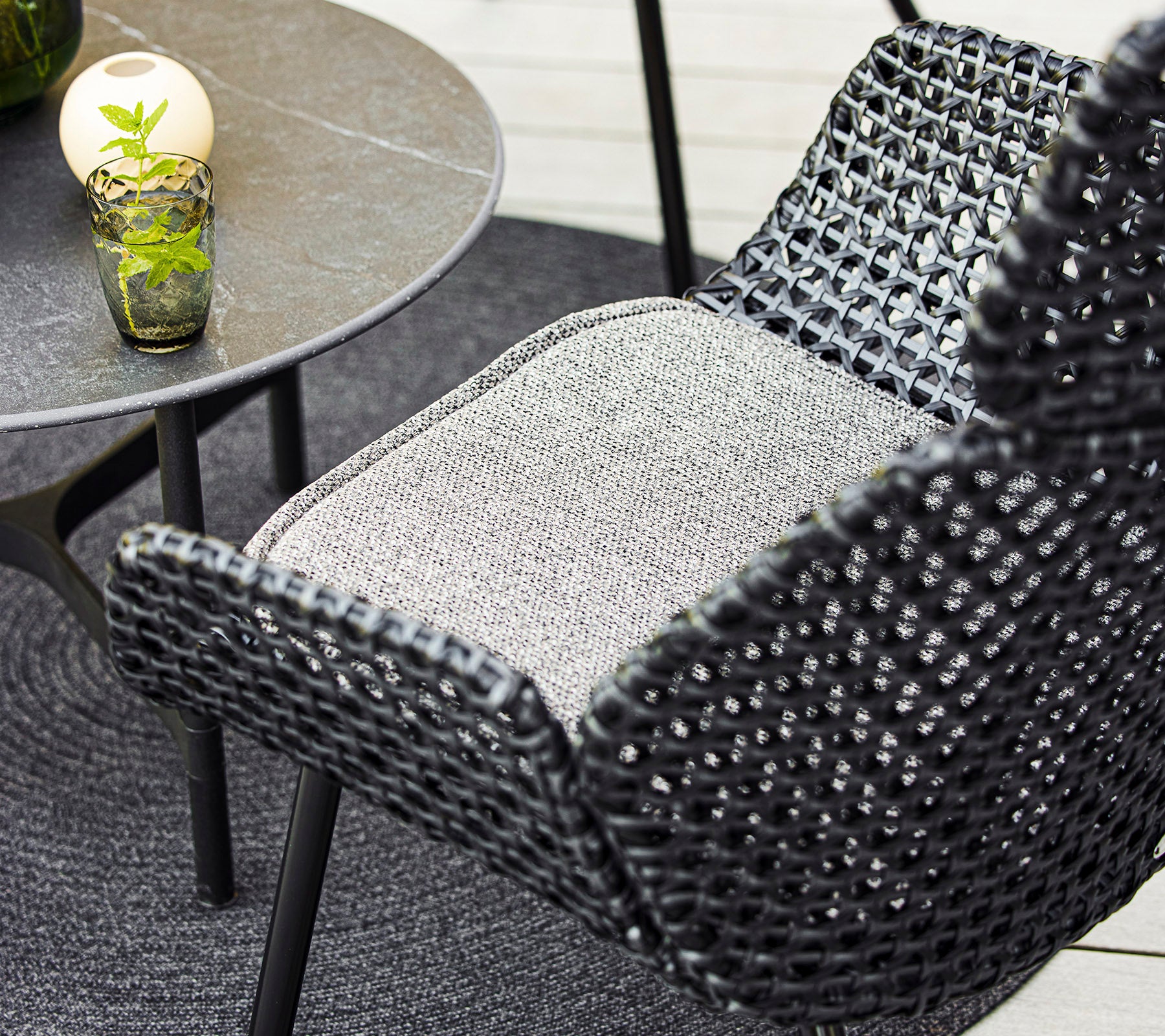 Een close-up van een stijlvolle gevlochten stoel en een klein tafeltje met een decoratief glas en een plant, gecombineerd met een gezellige ronde mat.
