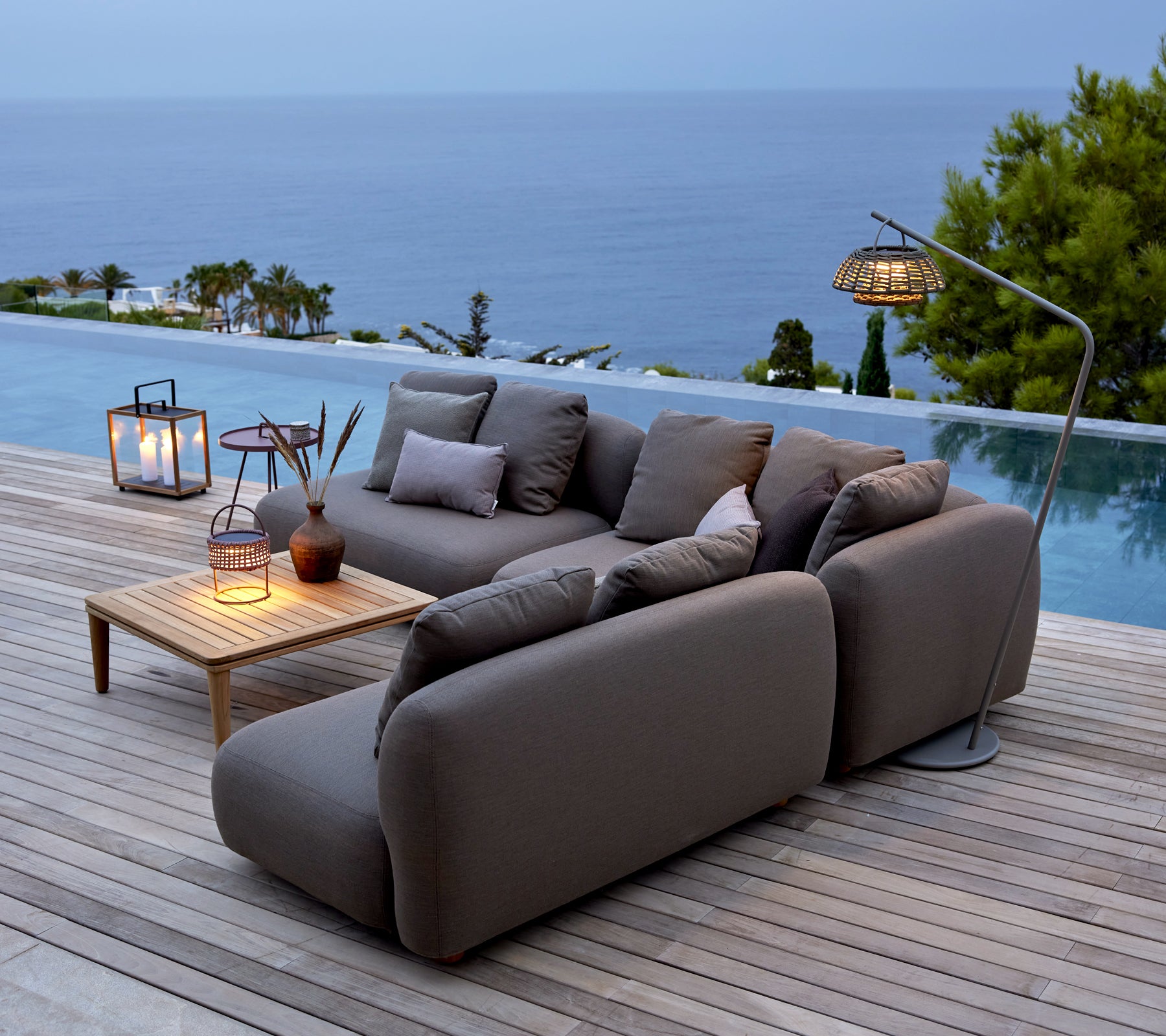 Grijze sofa met houten tafel en lantaarns bij een zwembad met uitzicht op de oceaan.