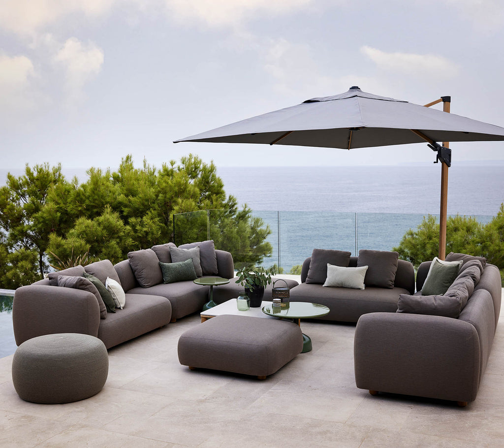 Modern lounge set met pluche kussens, vergezeld van een ronde tafel en een grote parasol, omgeven door groen en een schilderachtig uitzicht.
