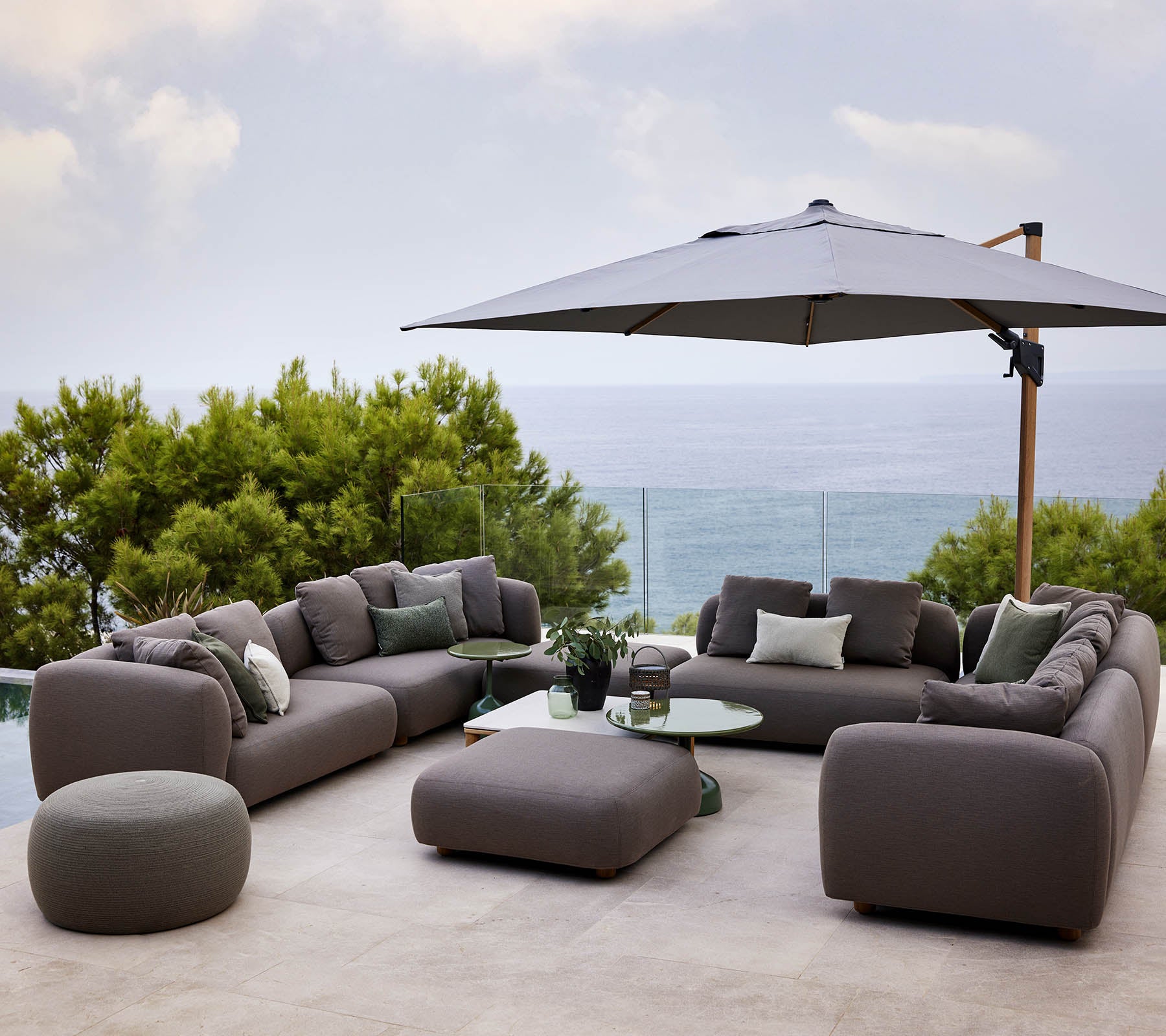 Modern lounge set met pluche kussens, vergezeld van een ronde tafel en een grote parasol, omgeven door groen en een schilderachtig uitzicht.