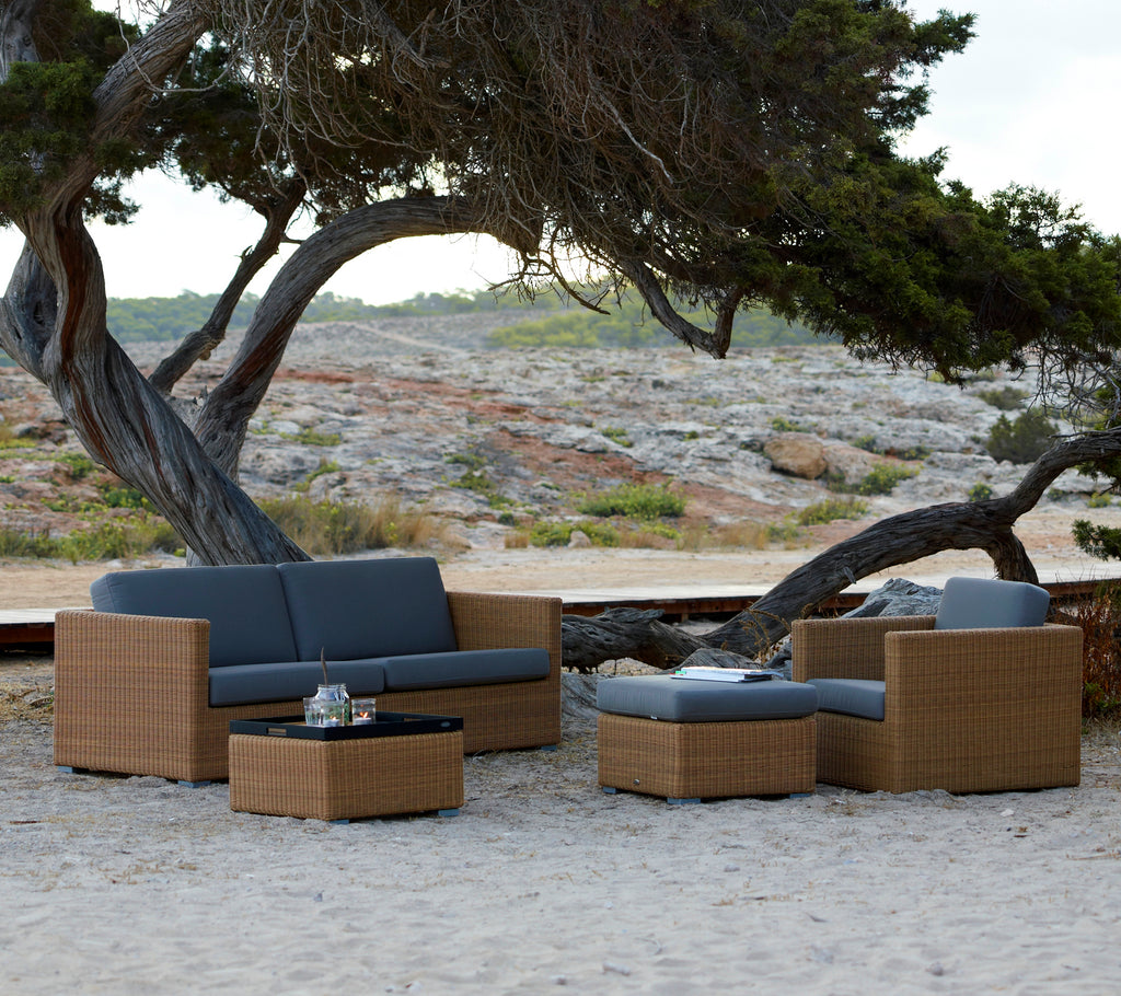 Moderne bruine lounge-meubels met grijze kussens, geplaatst in een natuurlijke buitenomgeving.