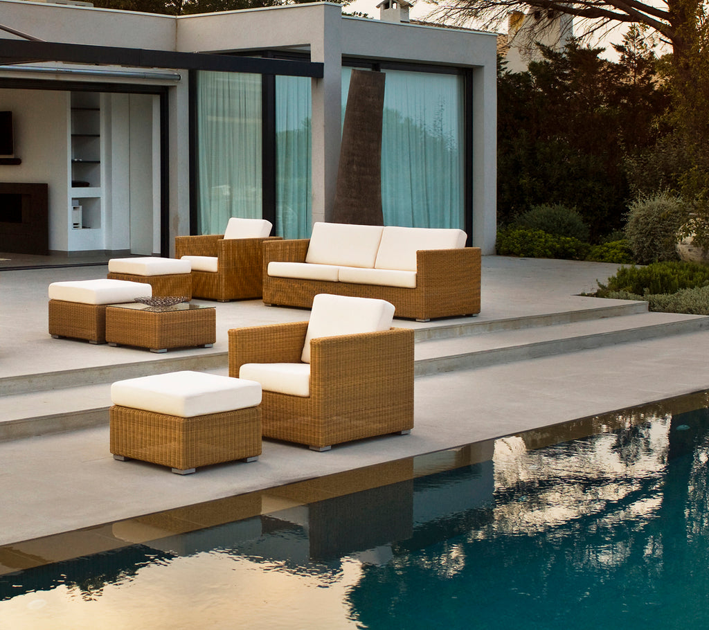 Moderne buitenlounge met een bruine bank, stoelen en kussens bij een zwembad.