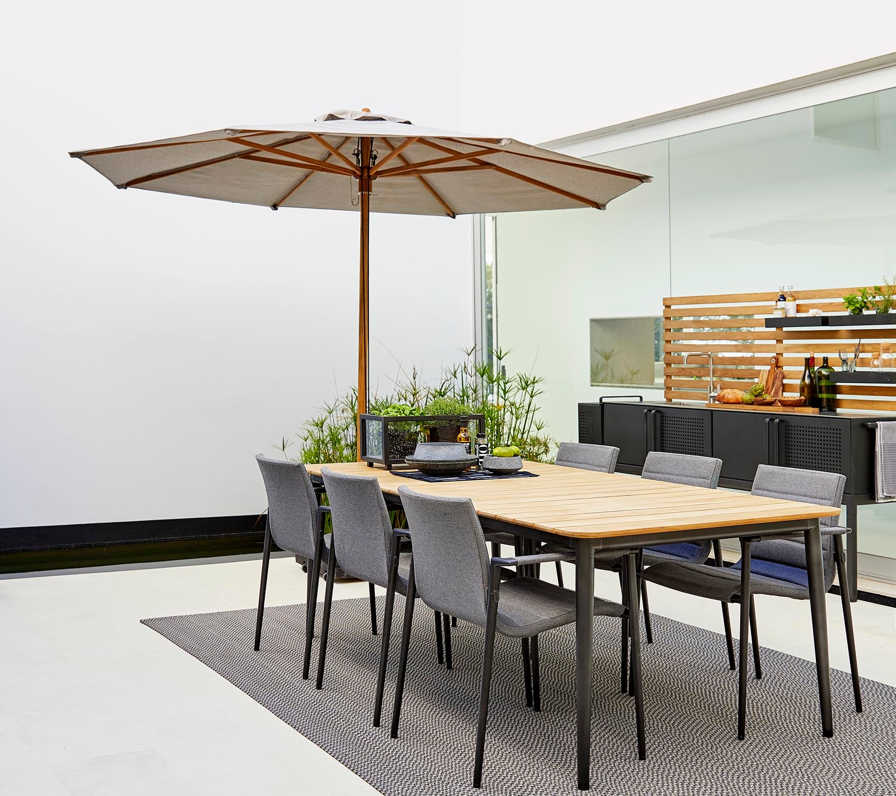 Een klassiek eetset met een houten tafel met een parasol en grijze stoelen, omringd door decoratieve planten op een stijlvolle vloerbedekking.