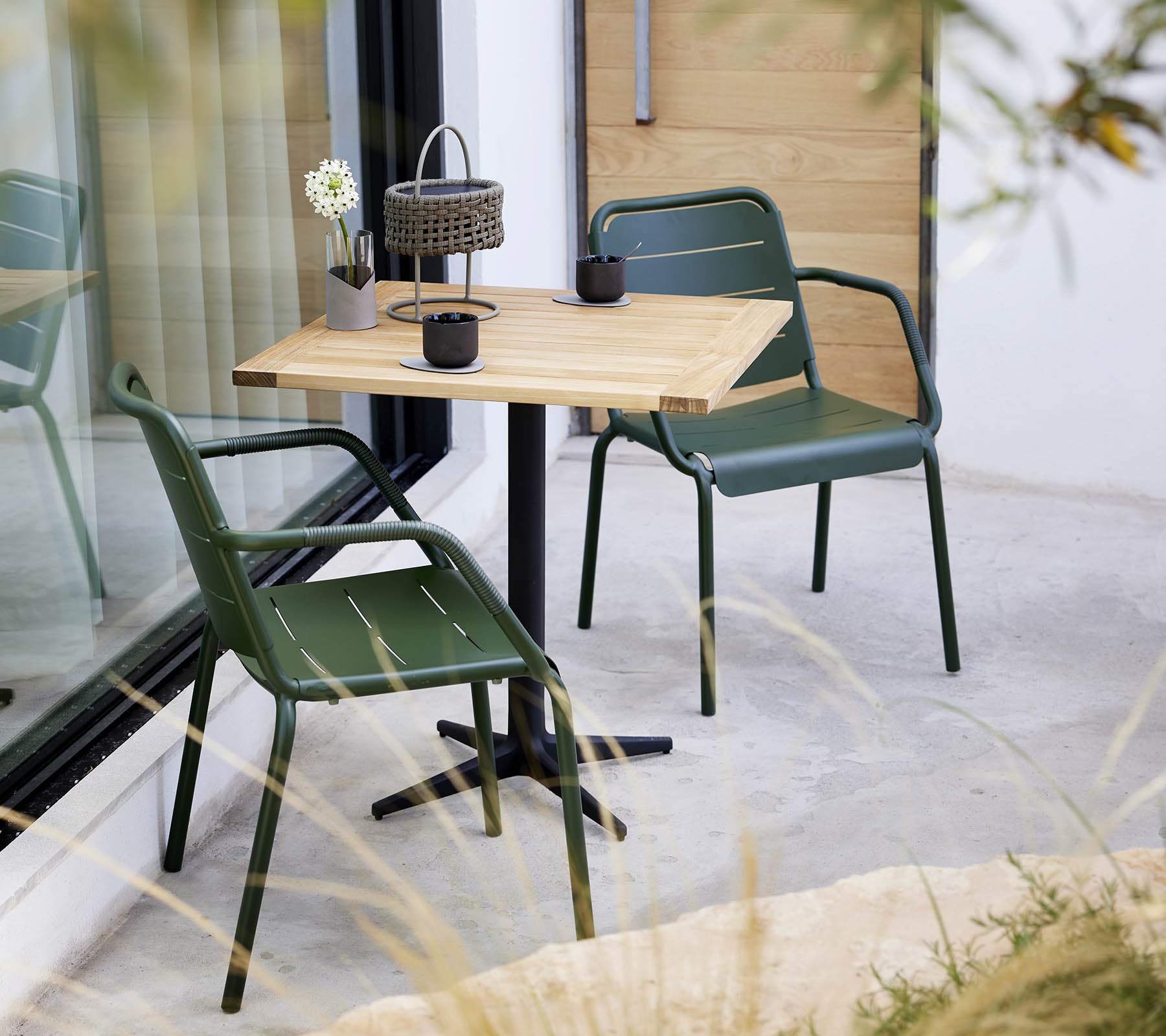 Een minimalistische setting met een houten tafel en groene stoelen, met een kleine bloemenarrangement en een decoratieve lantaarn.