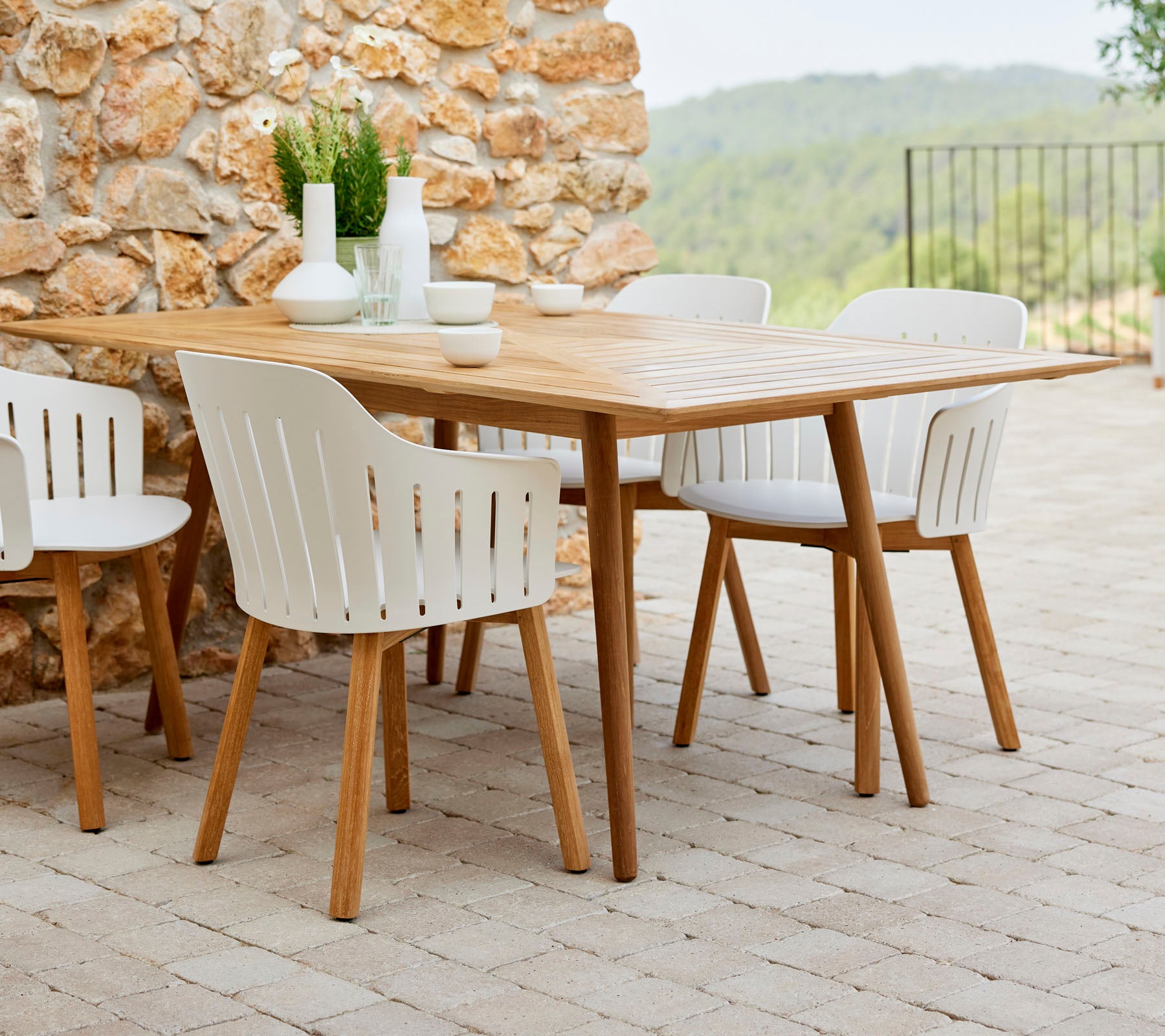 Een houten eettafel omringd door witte stoelen, aangevuld met decoratieve planten en eenvoudig servies, tegen een achtergrond van natuursteen.