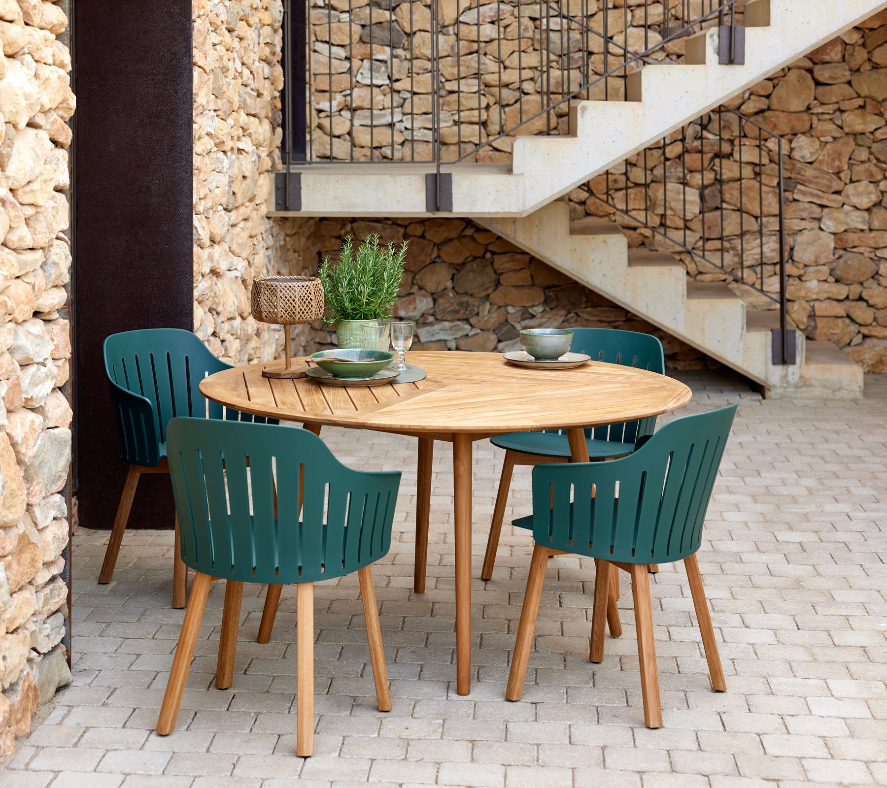 Een ronde houten tafel omringd door vier groene lattenstoelen, aangevuld met decoratieve kommen en een kleine plant.