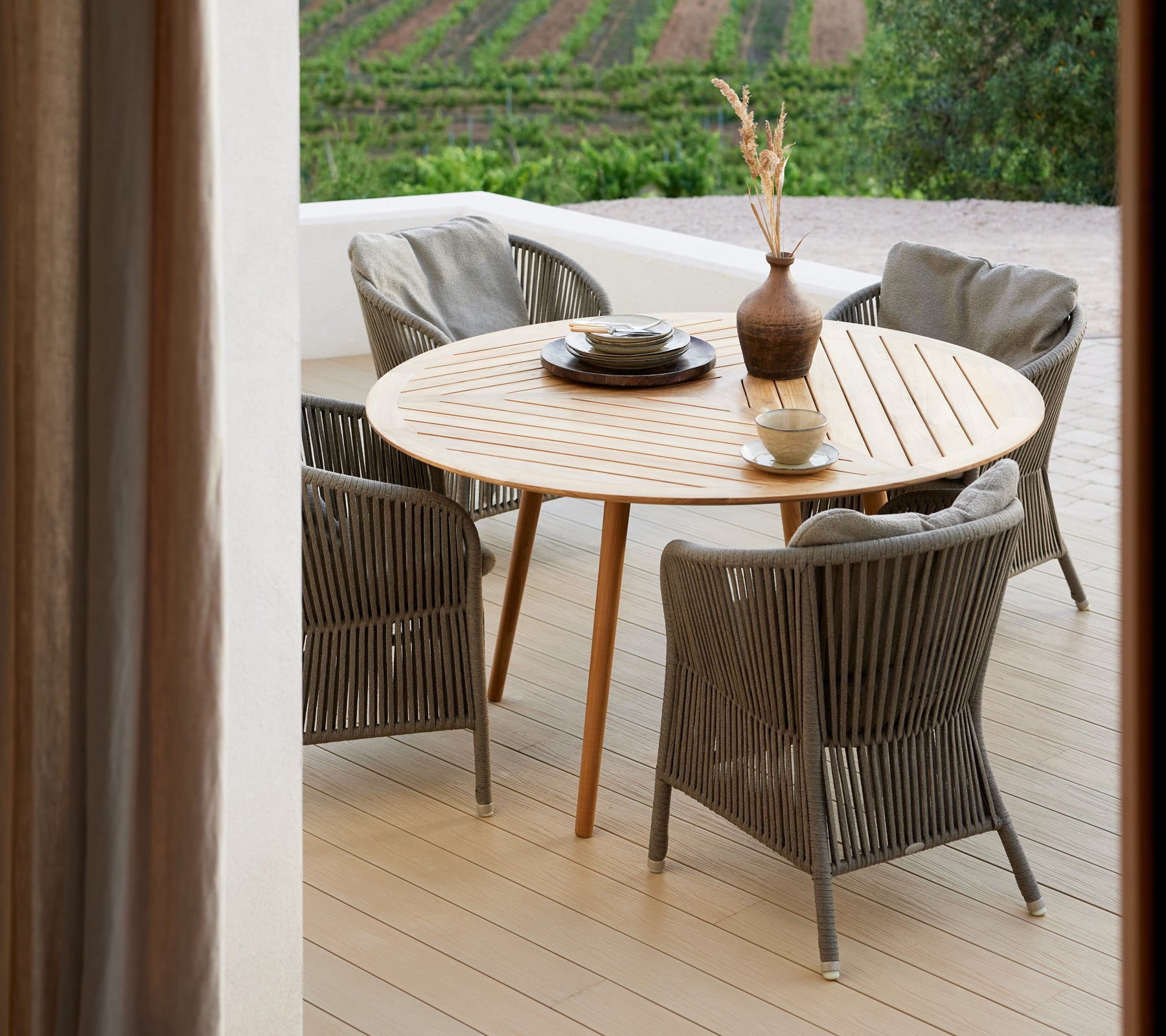 Een ronde houten tafel omringd door vier gevlochten stoelen, met een vaas en een decoratieve schaal, tegen een pittoresk decor.