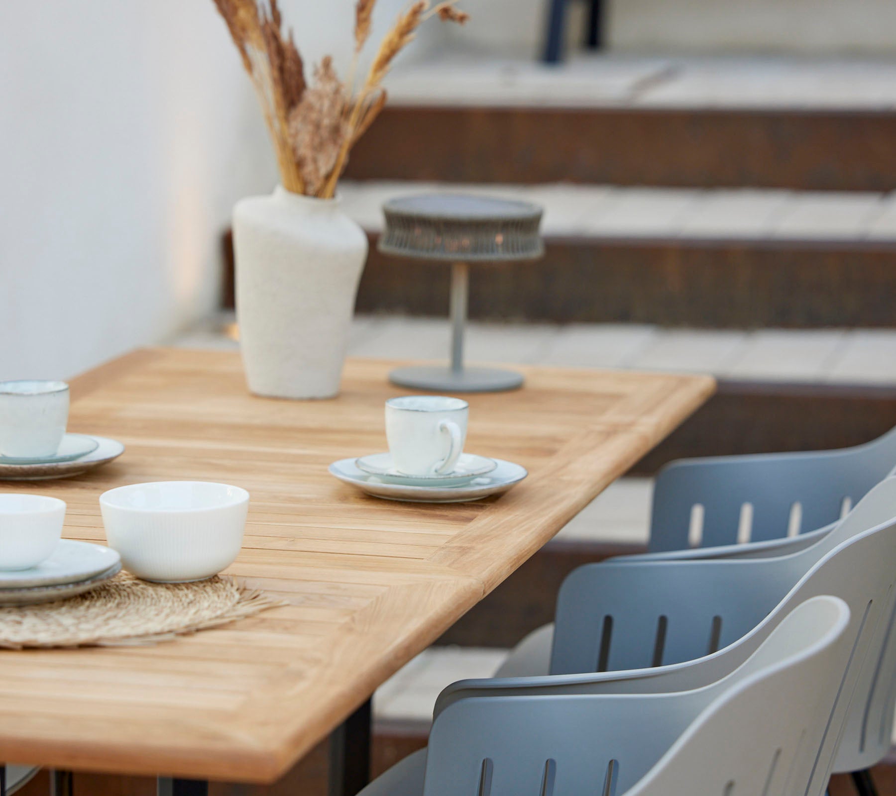 Een houten tafel gedekt met kopjes en borden, versierd met een vaas met gedroogde bloemen en grijze stoelen in de buurt.