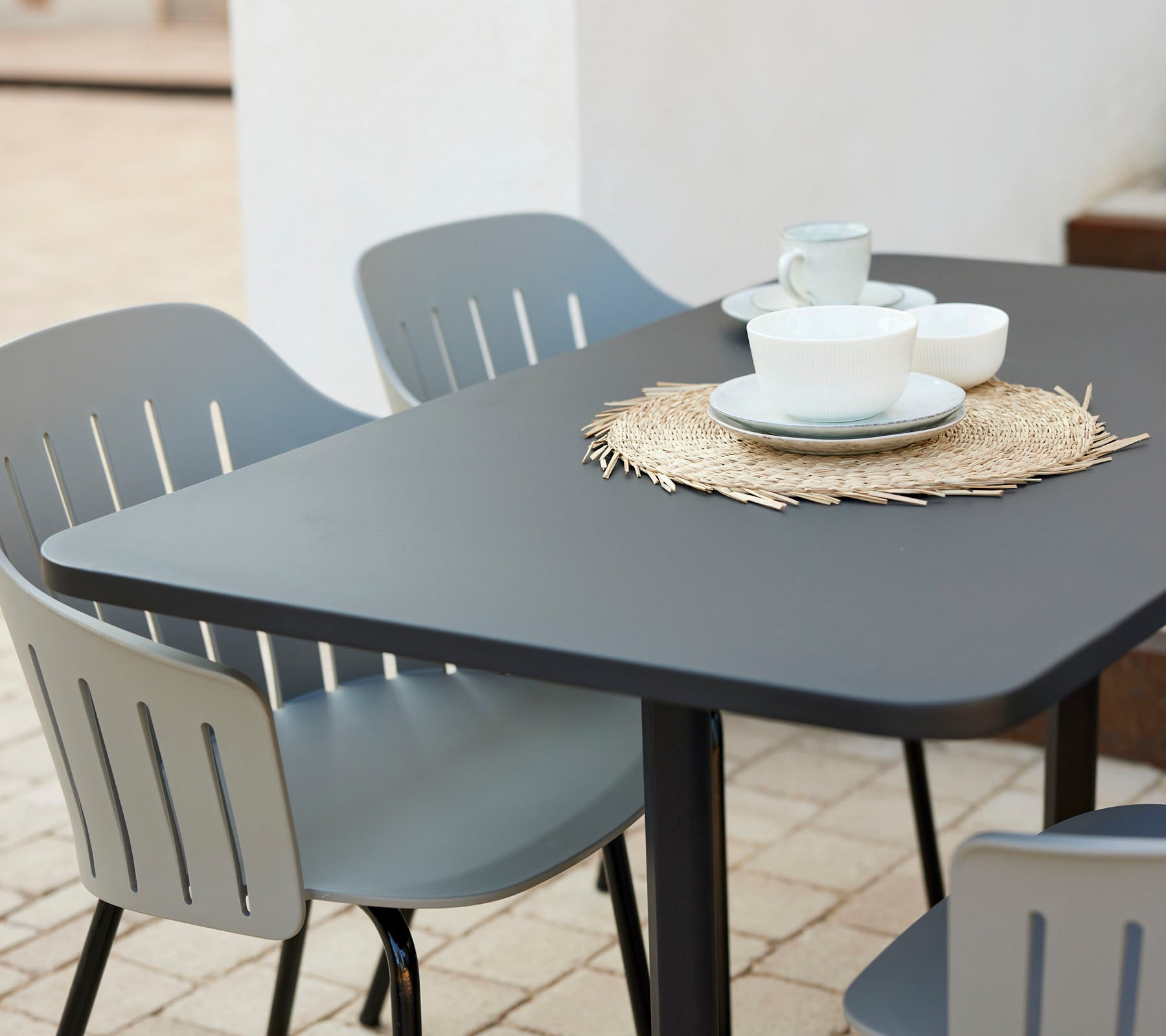Modern cafétafelset met een zwart tafelblad, omringd door grijze stoelen, met borden en kopjes op een rieten placemat.