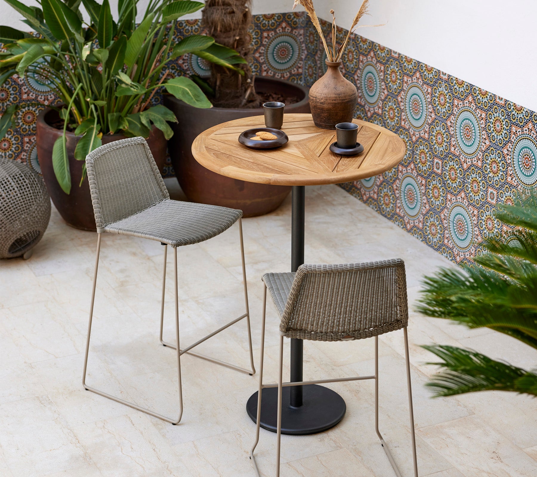 Een stijlvol bistroset met een ronde houten tafel en twee gevlochte krukken, vergezeld van decoratieve planten en modern servies.